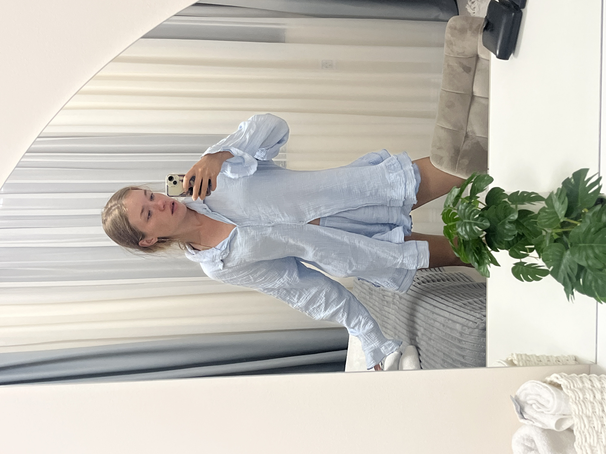 A young woman with blonde hair pulled back in a bun is standing in front of a mirror in what appears to be a bedroom or dressing area. She is wearing a light blue, crinkled fabric pajama set consisting of a button-down top with long sleeves and ruffled cuffs, and matching shorts with a ruffled hem. She is holding a smartphone in her right hand, taking a selfie, and looking down at the screen with a neutral expression.
The background is softly lit and consists of sheer curtains in shades of white and grey. To the left, a portion of a bed with a grey textured comforter is visible, along with some neatly folded white towels. To the right, a plush, tufted chair and a white tabletop are partially visible. In the foreground, on the tabletop, there is a woven basket containing rolled white towels and a potted plant with large green leaves. The overall scene suggests a relaxed, domestic setting, possibly in a hotel room or apartment in Ulcinj, Montenegro.