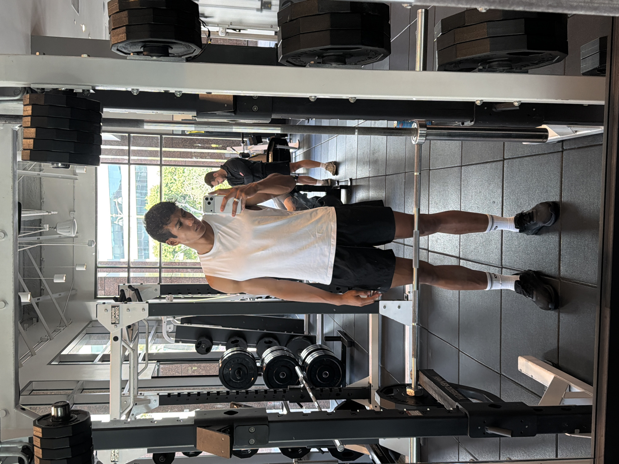 A young man stands in the center of a gym, holding a smartphone, presumably taking a photo or video of himself. He is wearing a white tank top, black shorts, white socks, and black sneakers. The gym appears to be well-equipped with weightlifting machines and free weights. Another individual is visible in the background, engaged in exercise. The gym has large windows that provide natural light and a view of a cityscape, potentially indicating an urban setting. Based on the architectural style visible through the windows, the location might be Los Angeles, California. The scene overall suggests a modern, health-conscious environment.