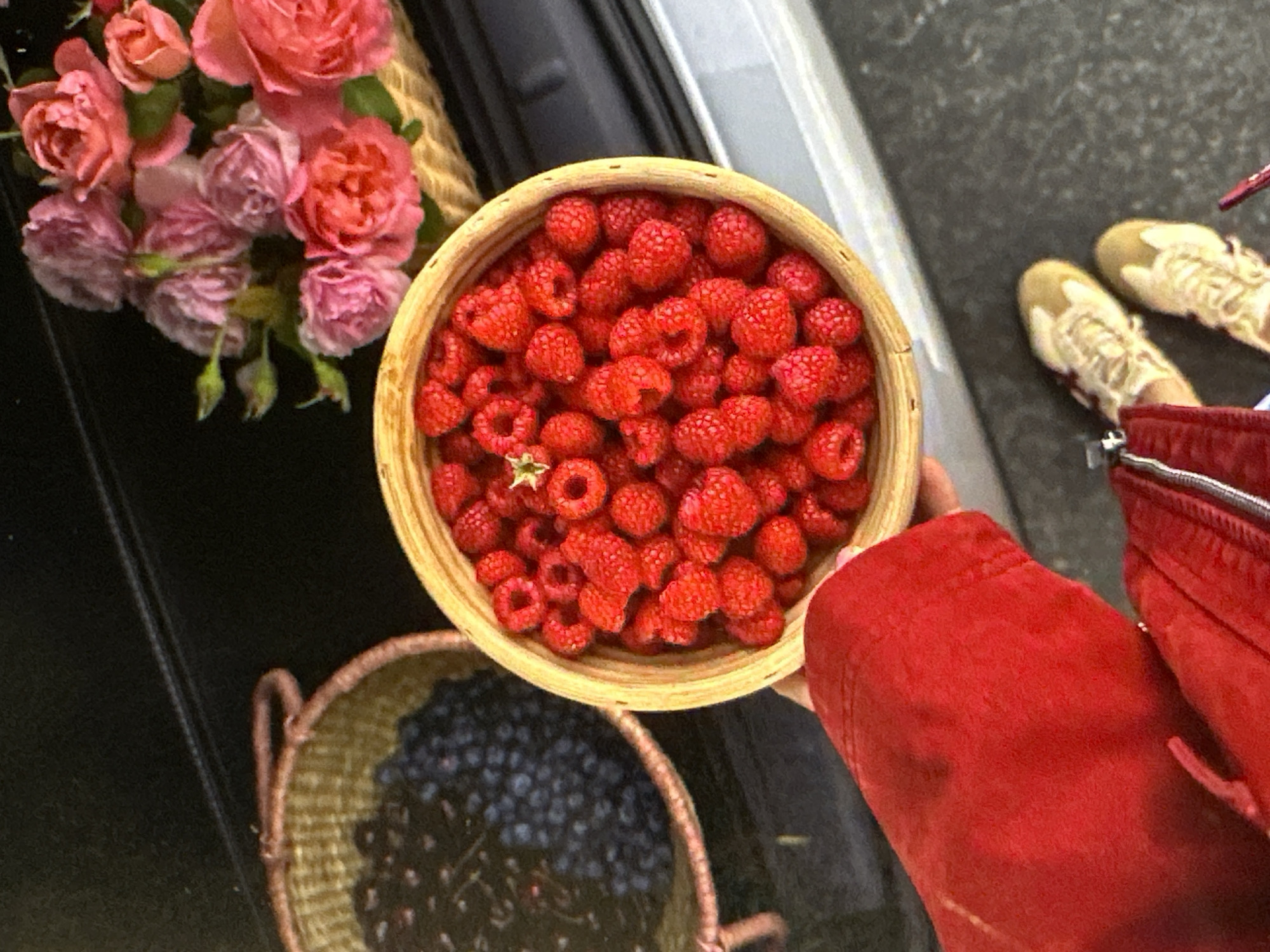 A high-angle shot presents a selection of fresh produce and flowers, potentially taken at a market in Yerevan, Armenia. Dominating the scene is a round, light-colored wooden bowl filled to the brim with bright red raspberries. Adjacent to this, a similarly shaped basket contains dark-colored blueberries or cherries. Beside these, a smaller basket is filled with pink roses.
A person wearing a red jacket is holding the bowl of raspberries. The edges of beige shoes are visible to the upper right. The scene is set against a mixed background of gray and black, possibly the pavement of a city street or the surface of a vehicle.