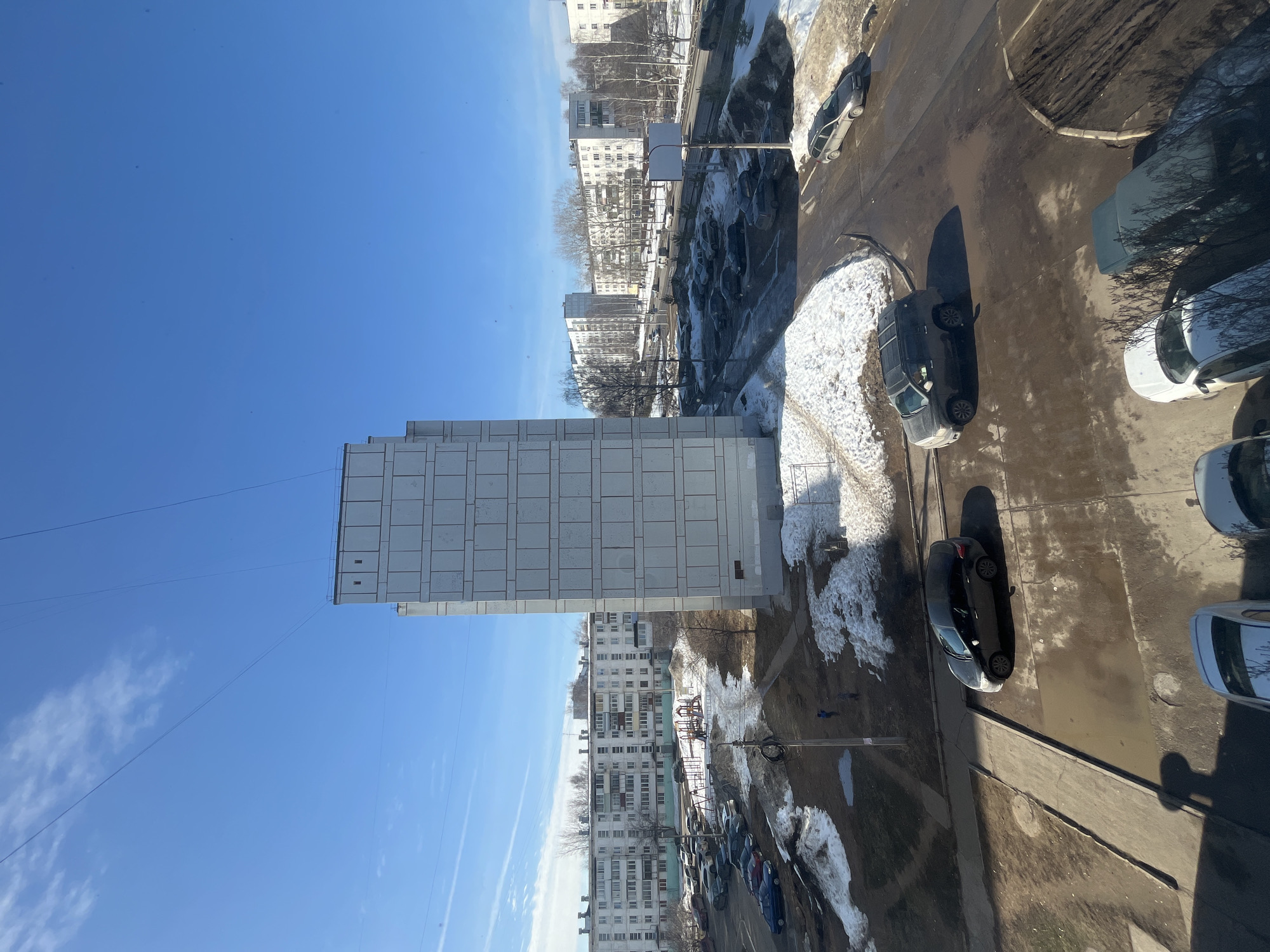 An elevated, wide-angle photograph depicts a residential district in Chaikovsky, Russia. The scene is illuminated by strong daylight under a clear blue sky with faint wispy clouds and overhead utility lines.
In the mid-ground, a prominent, tall, light-grey, multi-story residential building with a distinct grid-patterned facade stands centrally. To its left, a children's playground with colorful structures is situated on an irregularly shaped area of melting snow, exposed earth, and puddles. Two small figures, consistent with children, are present near the playground equipment. Further left and in the background, a long, white, multi-story apartment building with numerous windows and balconies extends horizontally, typical of Soviet-era architecture.
The ground throughout the scene displays significant patches of melting snow, exposed dark brown earth, and puddles on both paved asphalt roads and concrete pathways. Numerous vehicles, predominantly sedans and SUVs of various colors, are parked in several parking lots and along roadways. In the immediate foreground, a dark-colored sedan and a dark SUV are parked on a paved surface, partially obscured. A dark grey station wagon is parked on a muddy shoulder in the mid-right.
Bare deciduous trees are sparsely distributed among the buildings and parking areas. In the further background, additional multi-story residential buildings are visible, interspersed with more bare trees. Distinct shadows are cast by buildings and vehicles, indicating bright sunlight.