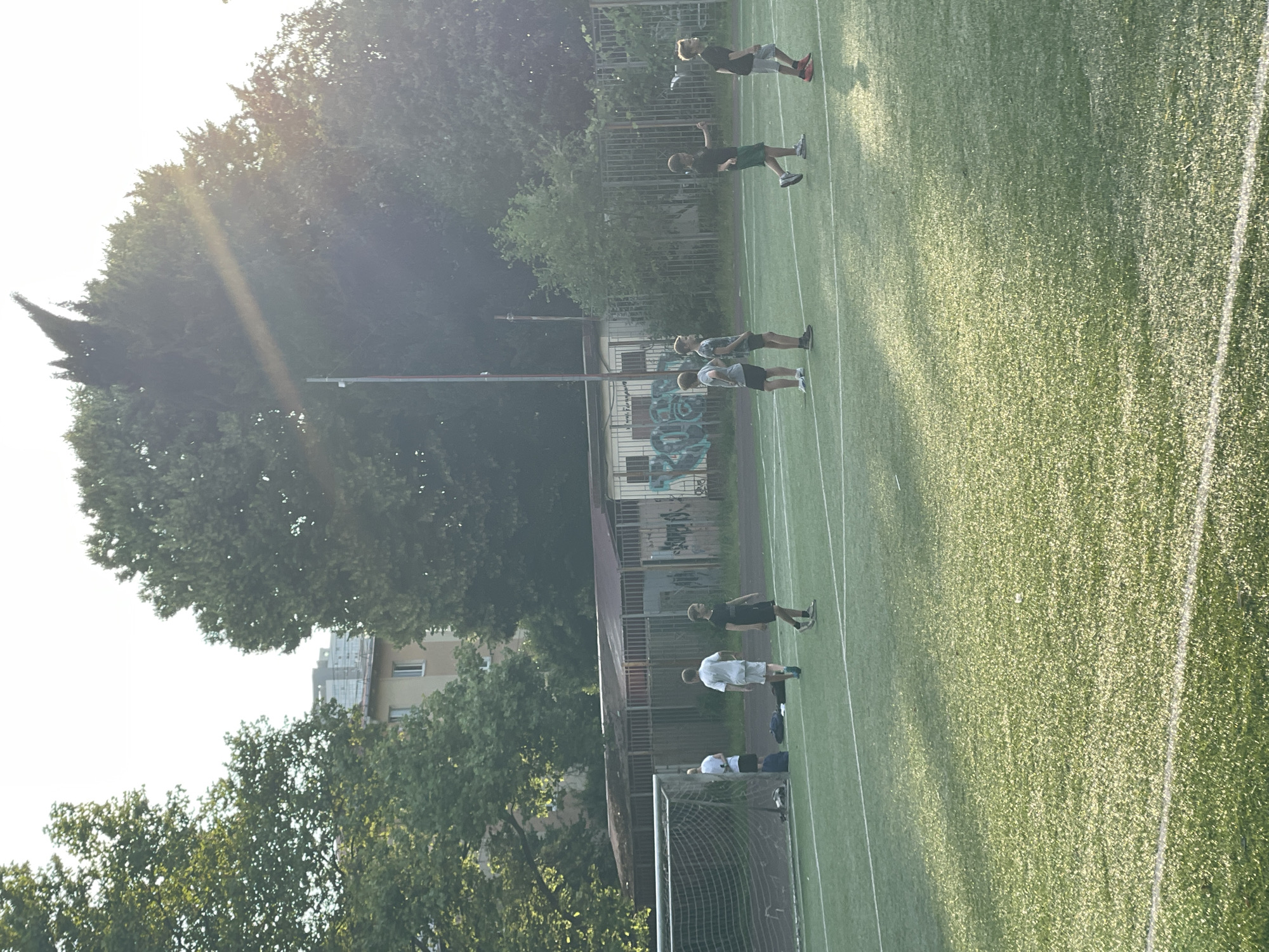 The image shows a group of young men on a green artificial turf field, likely a sports field. The field is marked with white lines, characteristic of a soccer or football pitch. Some of the men are standing in various formations, while others are in motion, suggesting a practice or training session. One person is lying on the ground near a soccer goal.
In the background, there is a fence, some trees, and buildings with graffiti visible on one of them. The lighting suggests it is either early morning or late afternoon, with sunlight casting long shadows and creating a warm glow on the field. The sky is bright, indicating clear weather.
The subjects are dressed in casual athletic wear, including t-shirts and shorts. The overall mood appears to be one of focused activity, typical of a sports team training. There is no discernible text in the image. The scene is set in an outdoor urban or suburban environment, with the presence of trees and buildings suggesting a location with some greenery and infrastructure.