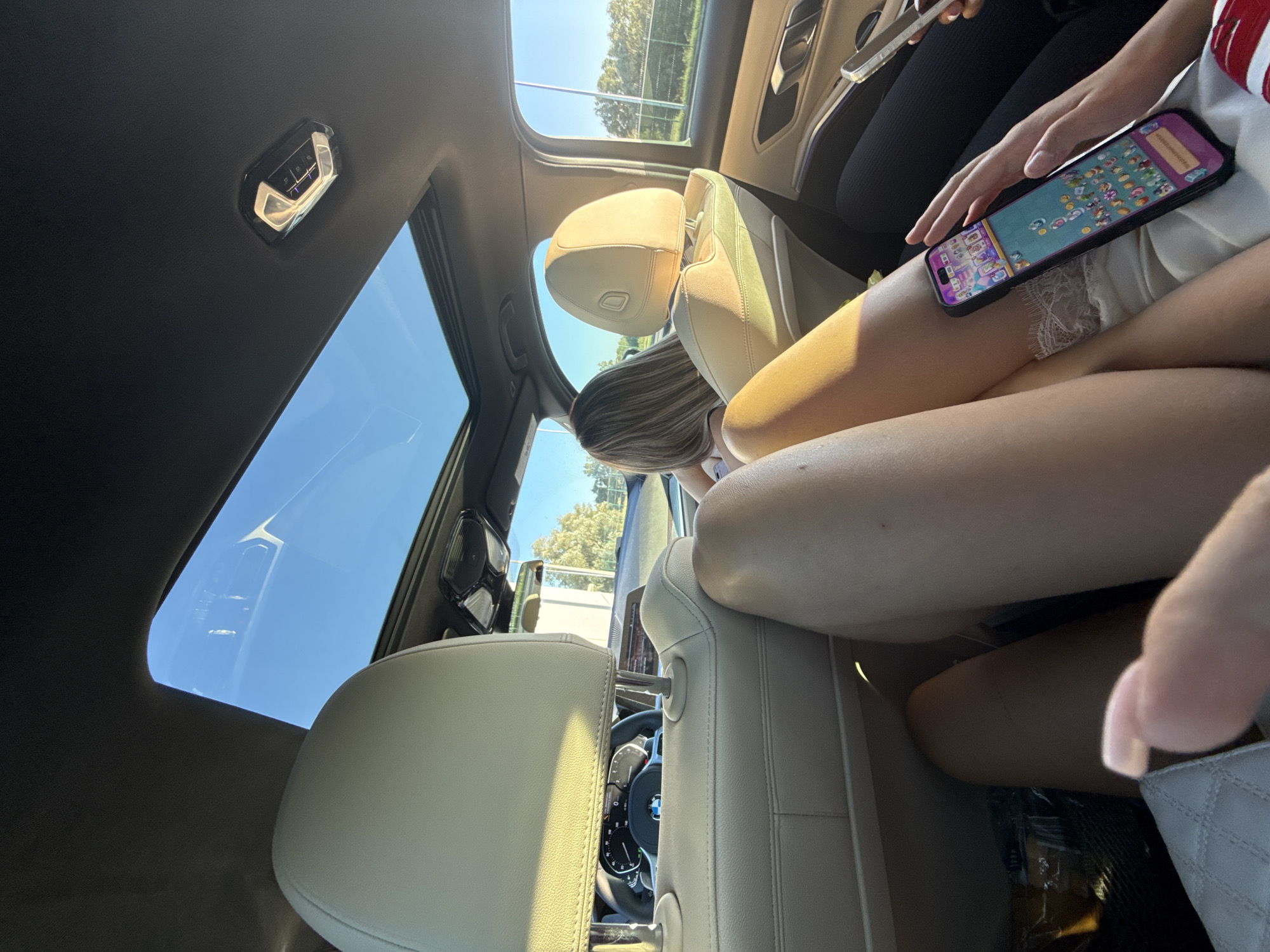 The image, taken inside a car looking towards the front and right, shows the interior of a modern vehicle with a panoramic sunroof open to a bright blue sky. The seats are light-colored leather. In the foreground, two individuals are seated, with their legs visible. One person, on the left, is partially obscured and their hand with a pink-tipped fingernail is reaching towards the viewer. The other person, to the right, is holding a smartphone displaying a colorful puzzle game, with their hand resting on their lap. The car's dashboard and steering wheel, featuring a BMW logo, are visible in the background. A woman with blonde hair is seen driving. Outside the car, through the windows, trees and a fence can be seen, suggesting an outdoor, possibly suburban or rural, environment. There is no specific indication of the city of Leova, Moldova in the image.