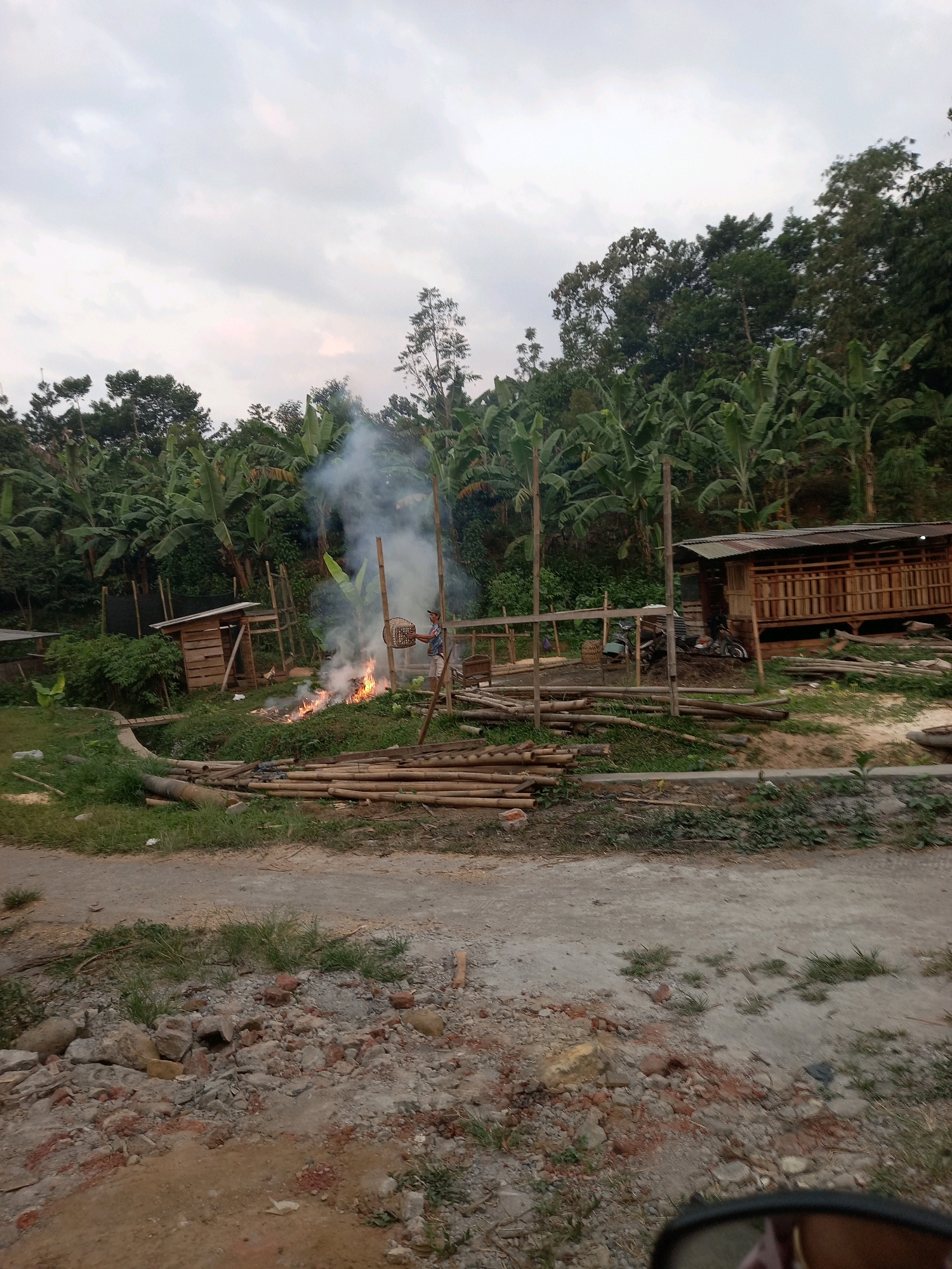 The image depicts an outdoor scene in a rural or semi-rural area, possibly on the outskirts of Wonosari, Indonesia. The foreground shows a dirt road littered with rocks and debris. In the middle ground, a man is visible tending to a fire that is producing a significant amount of smoke. He is holding a woven basket. Surrounding the fire are piles of bamboo poles, some stacked and others appearing to be recently cut. There are several rudimentary wooden structures, one of which resembles a small shack or shelter, and another larger structure that looks like an open-sided animal enclosure or workshop, constructed with bamboo and corrugated metal roofing. Lush green vegetation, including banana trees, dominates the background, suggesting a tropical or subtropical climate. The sky is overcast, indicating either early morning or late afternoon. The scene suggests a work in progress or daily activities in a village setting, possibly related to construction or land clearing.