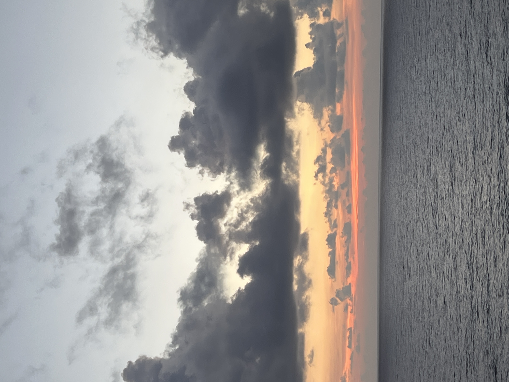 This is a photograph of the ocean at sunset. The sky is filled with dramatic clouds, ranging in color from light grey to dark, ominous shades. A strip of vibrant orange and pink hues can be seen just above the horizon, where the sun is setting. The ocean itself appears calm, with a gentle texture of small waves. The location is Unknown, Unknown. There are no people or man-made objects visible in the image.