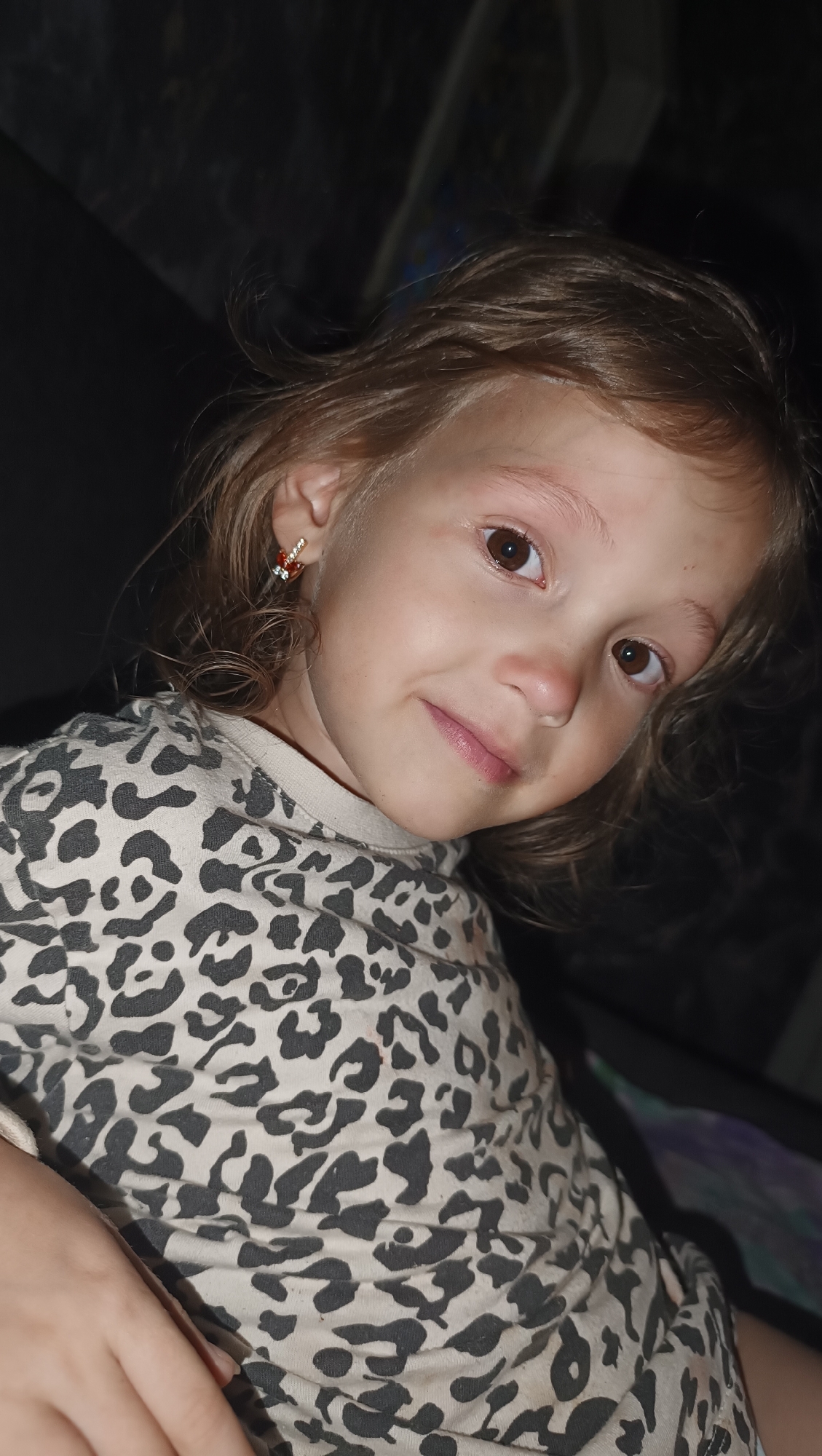 The image shows a close-up, portrait-style photograph of a young girl. She has light brown hair, brown eyes, and a fair complexion. She is looking slightly to her right, with a gentle smile on her face. The girl is wearing a light-colored shirt with a black leopard print pattern. She also has a small, decorative earring in her visible left ear, which appears to be gold with red and clear stones. The background of the image is dark and out of focus, making the girl the clear subject. There is no discernible information about the location, including any specific details about the city of Karsha, Russia. The scene is simple and focuses entirely on the child's facial expression and attire.