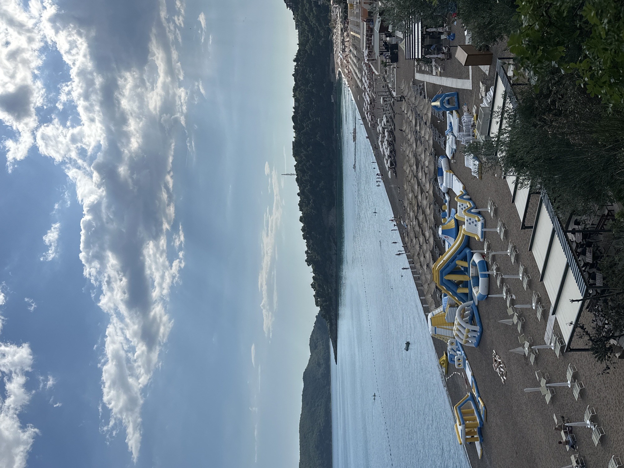 The image depicts a sunny beach in Boreti, Montenegro. In the foreground and right side of the image, are the beach amenities, including white sun umbrellas and sunbeds arranged in rows. Further along the beach, there are inflatable water play structures with yellow and blue colors. The beach has a sandy surface and is adjacent to the blue waters of the Adriatic Sea. A forested area extends along the left side, partially obscuring a tower structure. The sky is blue with a scattered layer of white clouds. People are seen on the beach and in the water.