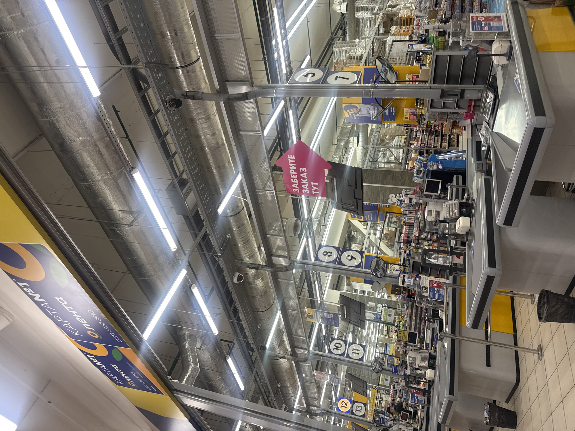 The image depicts the interior of a Lenta hypermarket in Saint Petersburg, Russia. The view is oriented along the checkout lanes, which are numbered from 5 upwards towards the background. The environment is brightly lit by rows of fluorescent lights suspended from the ceiling, along with extensive ductwork. The checkout lanes are modern, with electronic payment systems. Above the lanes are signs indicating the lane number. In one lane, a pink, arrow-shaped sign reads "ЗАБЕРИТЕ ЗАКАЗ ТУТ" (Pick up order here). Shelves stocked with a variety of goods line the aisles beyond the checkout areas. A yellow sign, reading "КАРТА ЛЕНТА" is visible along the left edge of the image.