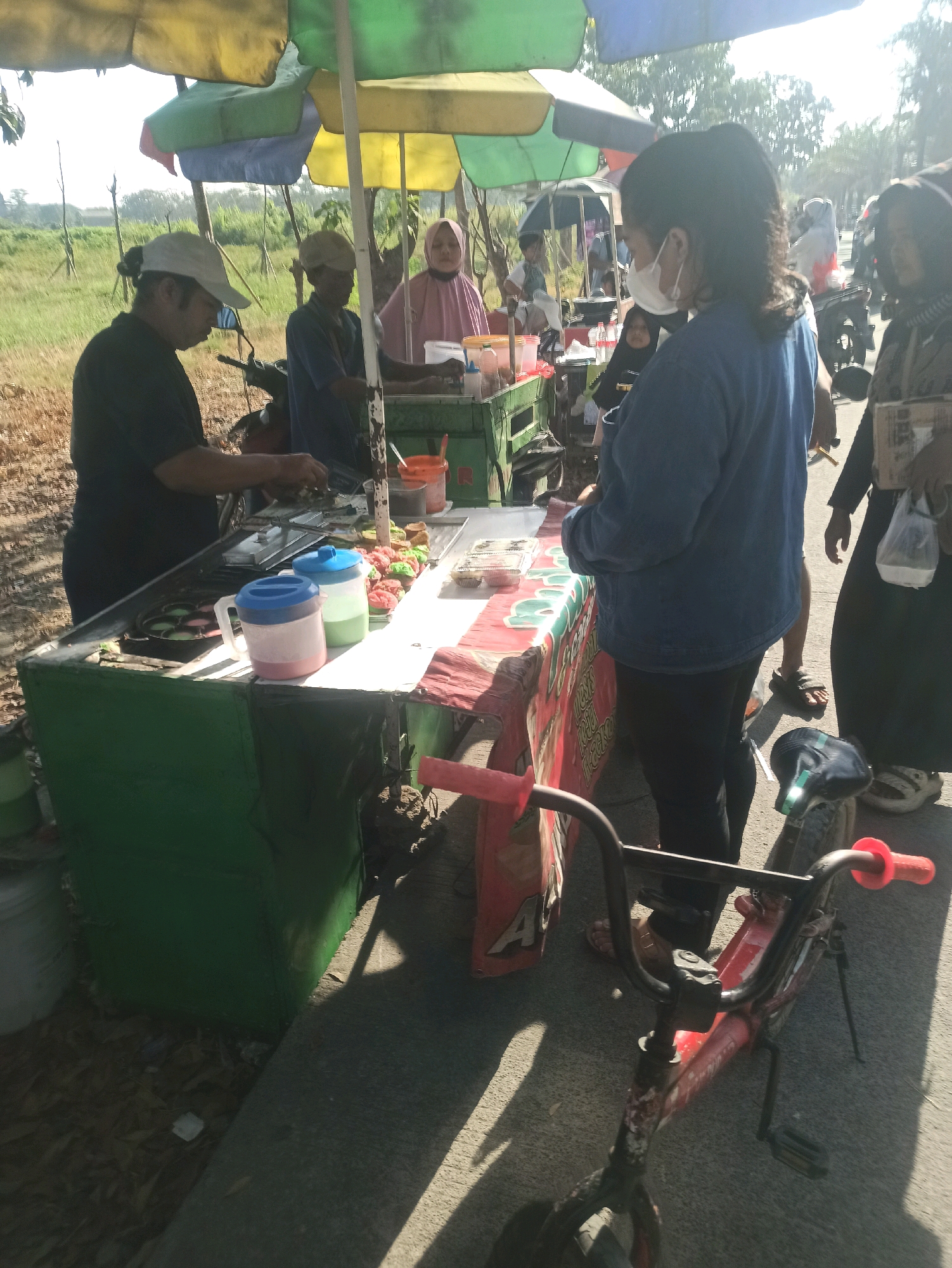 In Telagamurni, Indonesia, on a sunny day, an outdoor food stall is set up under colorful umbrellas. A man wearing a white cap and a dark t-shirt stands behind the stall, preparing food. To his right, another man in a blue shirt is also working. A woman in a pink hijab is visible further back. In the foreground, a woman wearing a blue denim jacket and a white face mask is interacting with the vendor. Next to her, another woman in dark clothing and a headscarf holds a plastic bag. A red bicycle is parked in front of the stall. The stall itself is decorated with a red and white banner. The surrounding environment appears to be an open area with vegetation and trees in the background, suggesting a roadside or market setting.