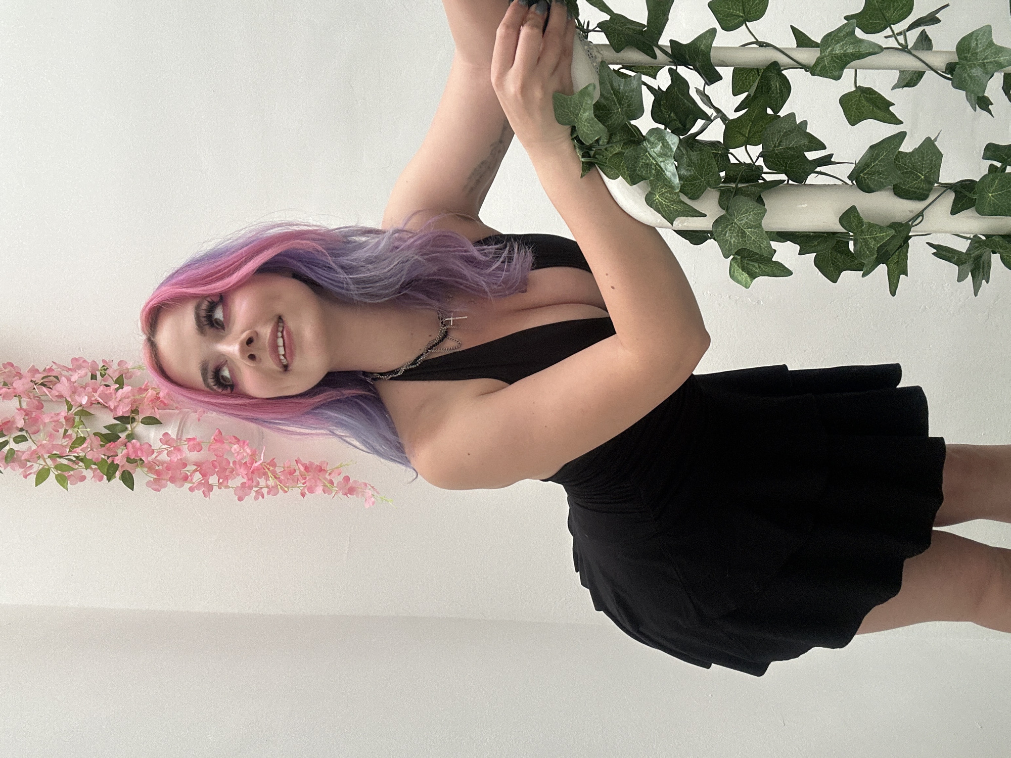 A young woman with striking long hair, colored in pink and purple ombre, is the central subject of this indoor portrait. She is wearing a black, deep V-neck mini-dress and a silver chain necklace with a cross pendant. Her hands, featuring dark nail polish, are holding onto a white pole or beam that is artfully entwined with artificial green ivy leaves. To her left, a decorative arrangement of artificial pink flowers adds a splash of color to the scene. The woman is smiling gently and looking directly towards the camera, her head tilted slightly, conveying a cheerful and engaging expression. Her eyes are enhanced with visible makeup, including eyeshadow and long eyelashes. The background consists of a plain white wall, indicating a brightly lit studio or minimalist interior setting. The image is clean and well-lit, with no visible text. While the image itself does not contain specific geographical markers, it is provided with the context of being from the City of Zagreb, Croatia.