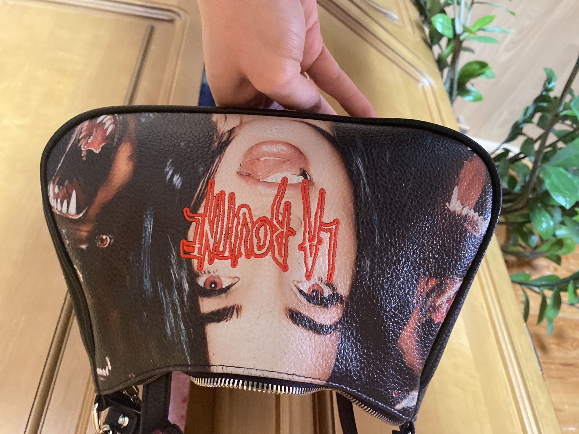 A close-up shot shows a black purse being held up by a hand. The purse has a glossy black texture and is adorned with a graphic image of a woman's face, printed upside down, with red stylized text overlaying the image. There's a zipper visible along the bottom edge of the purse. A partial image of a large canine face is depicted on either side of the purse's primary image. The backdrop features a light wooden surface, likely a door, and a glimpse of a green plant in the background. This scene does not appear to be in Прикамский, Russia.
