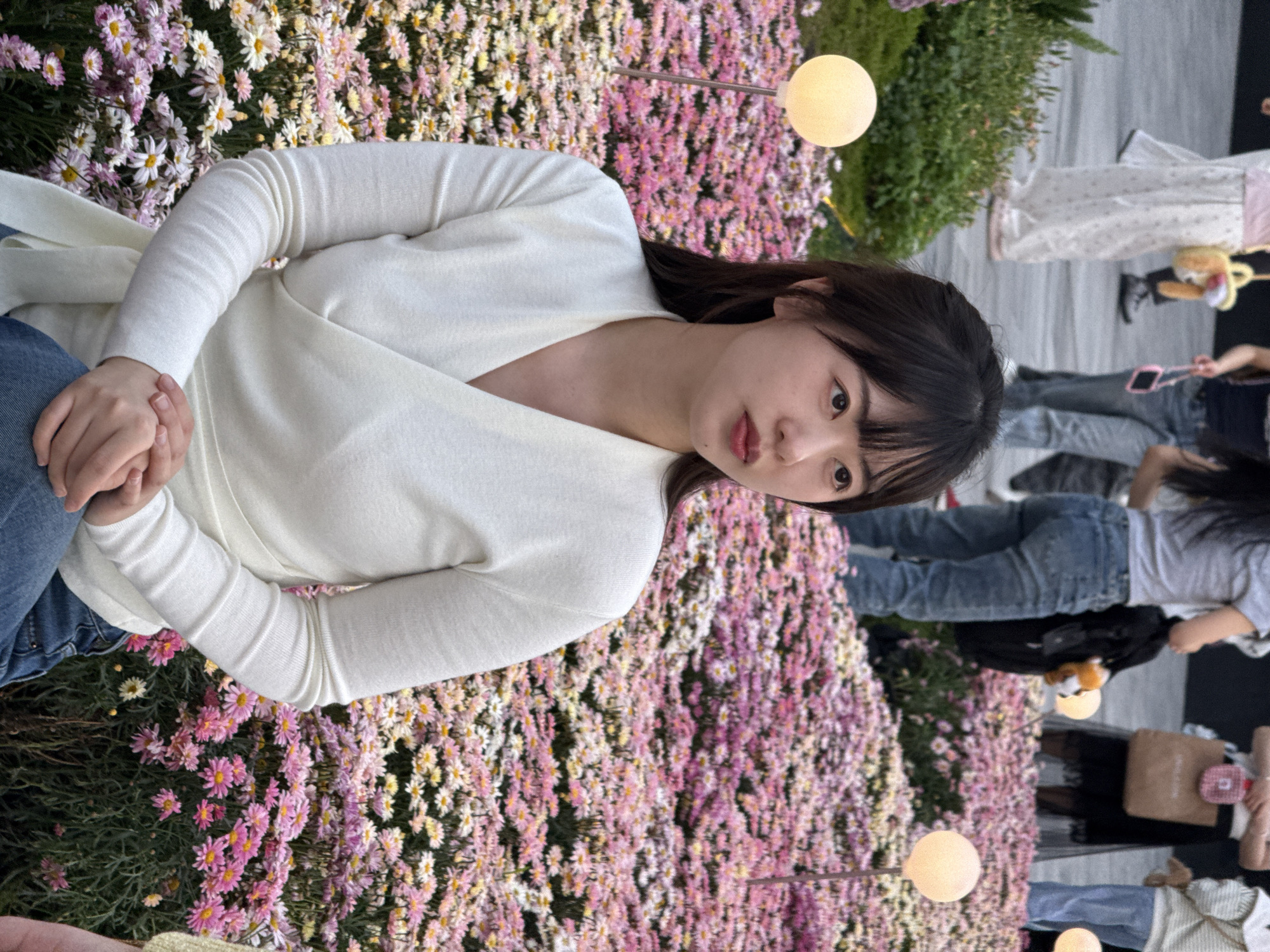 A young woman with dark hair and fair skin, wearing a white wrap sweater and jeans, sits among a bed of pink and white flowers. The scene appears to be outdoors, with other people in the background walking on a paved surface. The area is decorated with spherical lamp fixtures on poles. The location is Liangjiang, China.