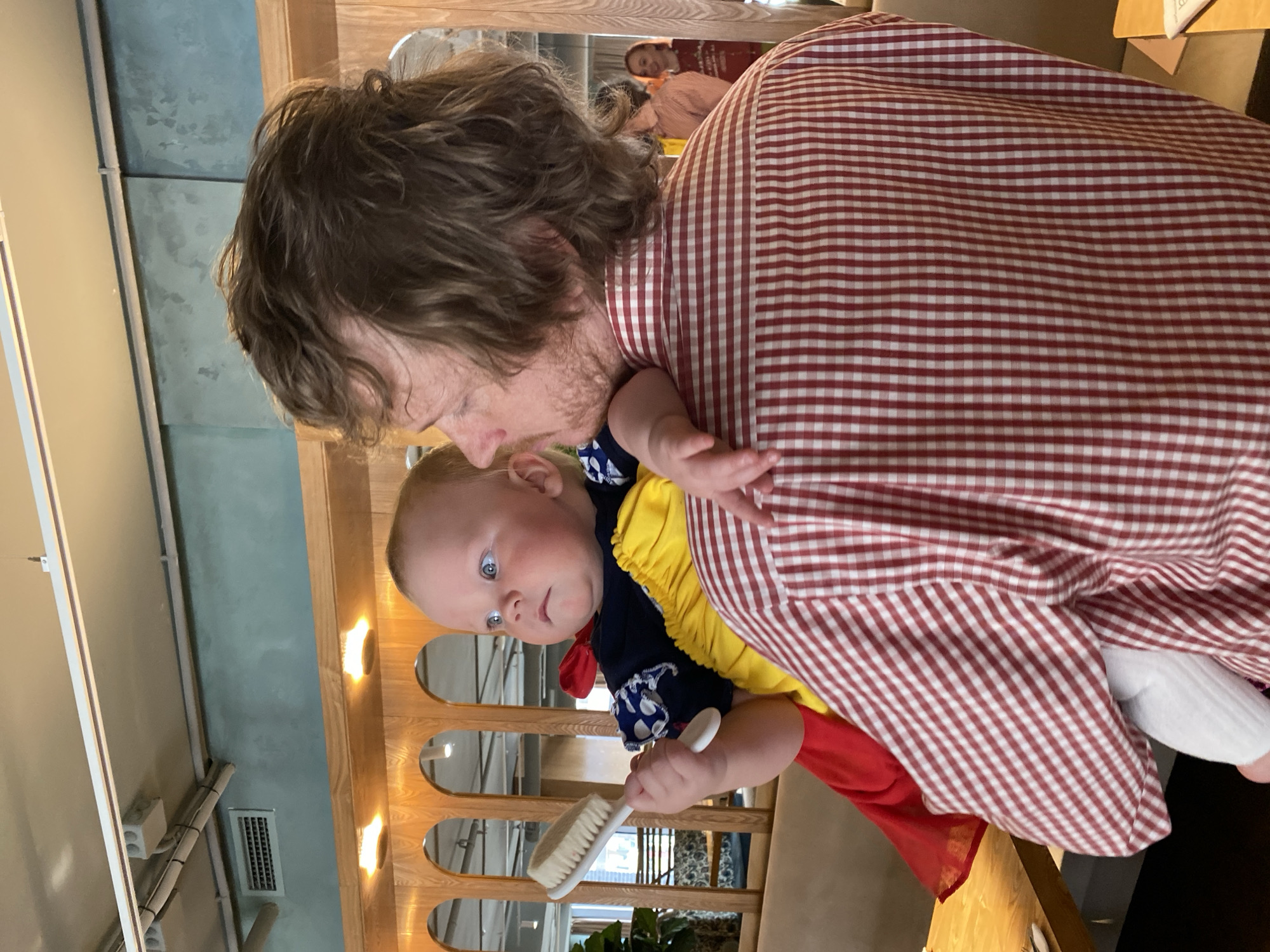 A man in a red and white checkered shirt is holding a baby. The baby has blue eyes and is wearing a yellow, blue and red outfit, and is holding a white brush. The man's head is near the baby's head as if he is kissing the baby. The photo appears to be taken inside a restaurant or cafe, with wooden decorative panels in the background, near a window with plants. It is impossible to verify if this scene is in Yekaterinburg, Russia.