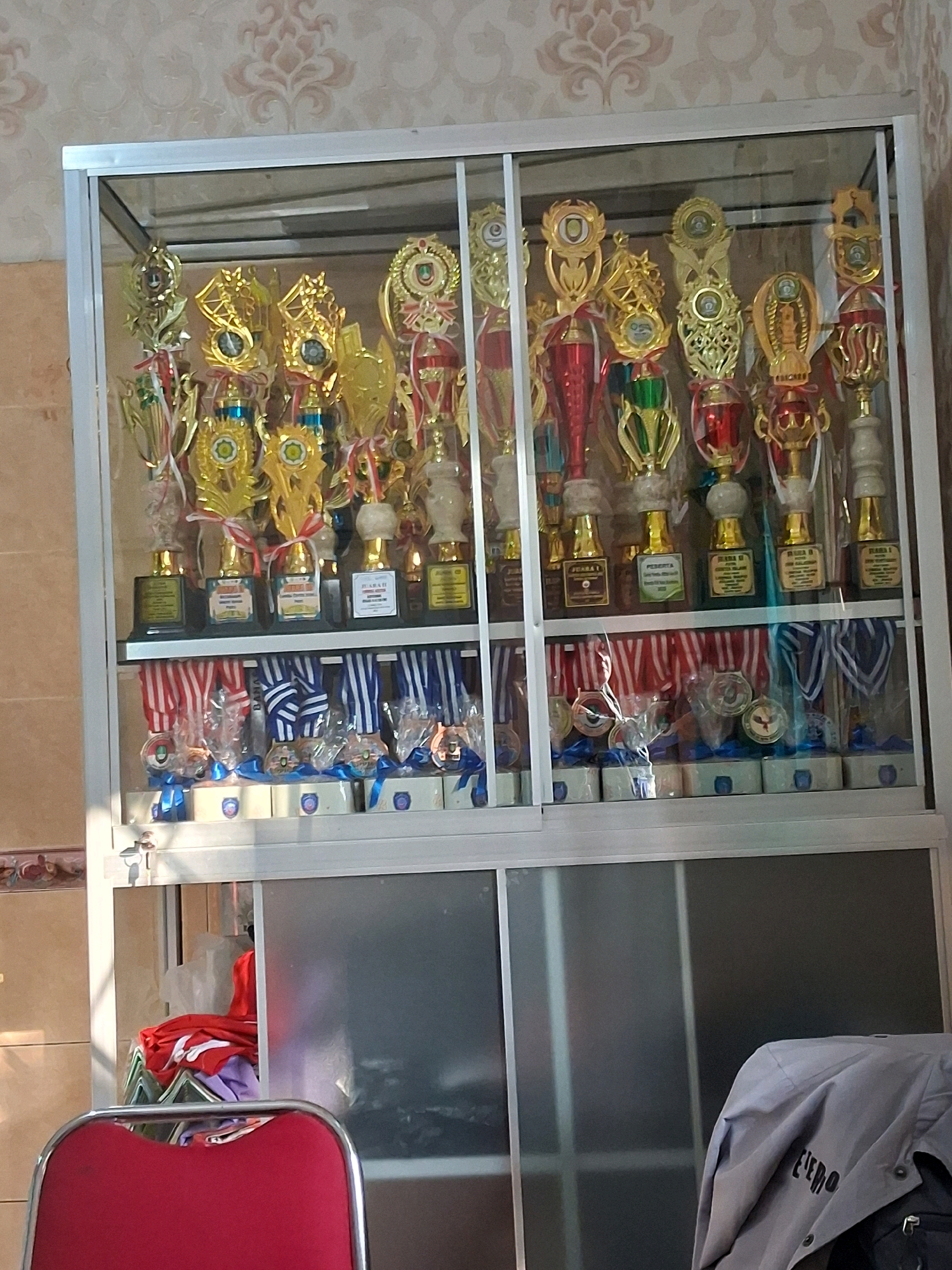 A glass-fronted display cabinet contains rows of trophies and medals. The top shelf holds numerous gold and red trophies of varying sizes, many with red ribbons. The bottom shelf displays an assortment of medals, each attached to a red and white or blue and white striped ribbon, and some resting on white boxes with blue ribbon accents. The cabinet is positioned against a patterned wallpaper. Below the cabinet, a red chair with a metal frame is partially visible. In the background of the lower cabinet section, a pile of folded clothing, including red and purple items, is seen. The scene appears to be indoors, possibly in a home or community center in Surakarta, Indonesia.