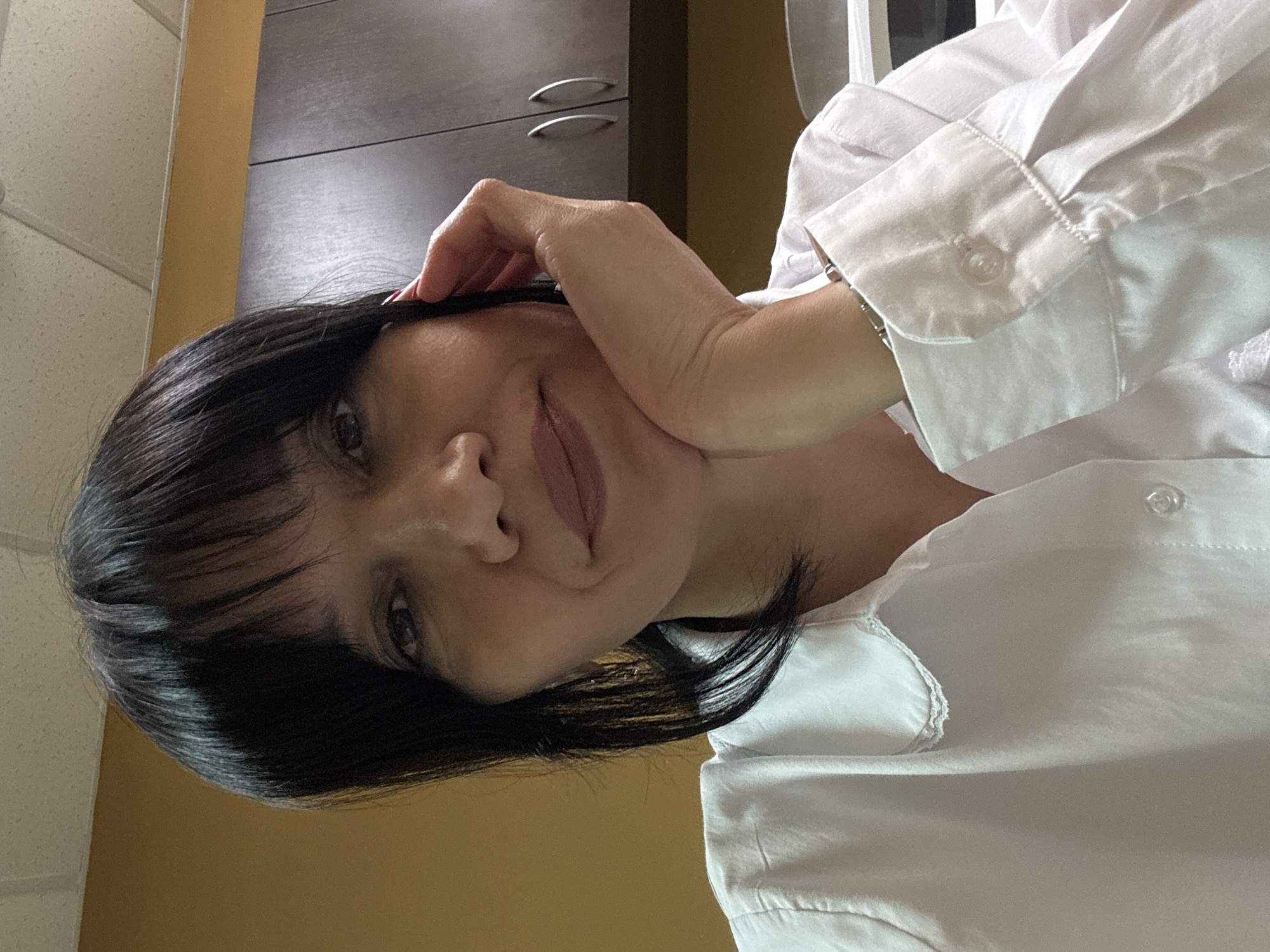 A woman with black hair and bangs is wearing a white, buttoned shirt. She is touching her forehead with her right hand, with her face angled towards the camera. Behind her are closed cabinets with silver handles and a wall that is partly beige and partly white with a textured pattern. The location is not discernible from the image; there's no evidence to suggest that it's in Chaikovsky, Russia.