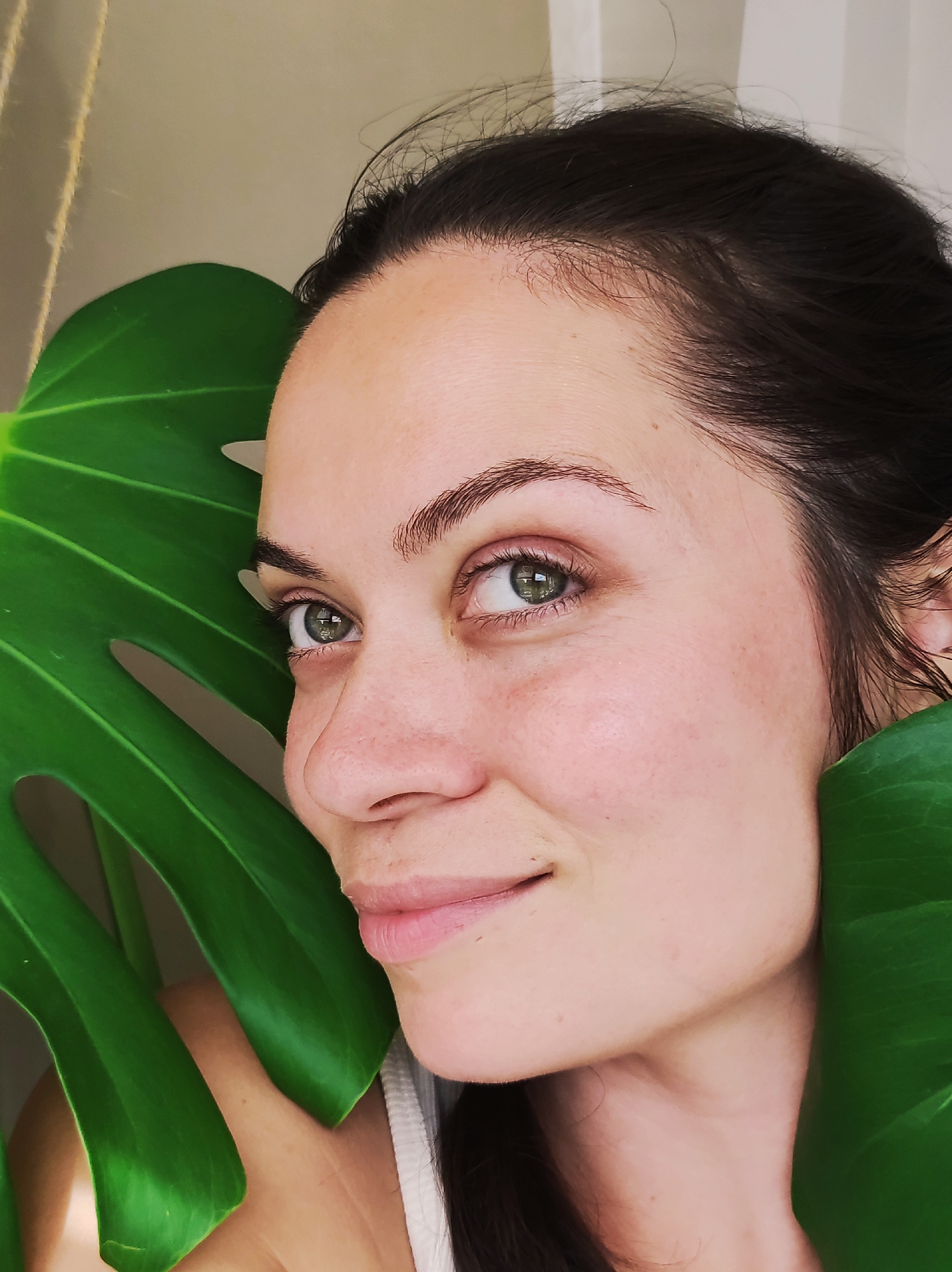 A close-up, head-and-shoulders portrait shows a woman with dark hair and green eyes, partially obscured by large, vibrant green leaves of a Monstera plant. She is looking to her left, with a gentle smile. Her skin appears fair with subtle freckles. The background is a soft, neutral tone, suggesting an indoor setting. The image does not contain information about the city of Chaikovsky, Russia.