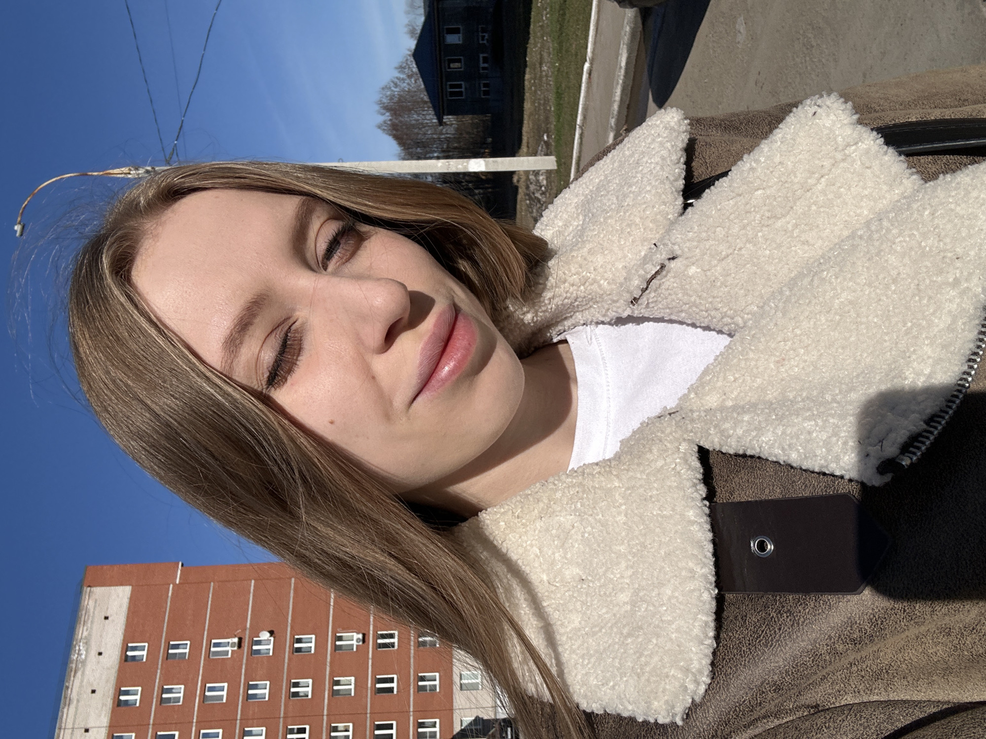 A young woman is the primary subject of a selfie taken outdoors. She has light brown hair and appears to be wearing a white t-shirt under a brown jacket with a cream-colored, fur-like trim around the collar. Her eyes are partially closed, and she is looking towards the camera.
The background of the image includes a clear blue sky and several buildings. There is a red brick building with numerous windows on the left side of the frame and a darker building with fewer visible windows further back. There also appears to be a power line crossing the upper left portion of the sky.
Based on the limited information, it is impossible to confirm the exact location within the city of Chaikovsky, Russia, without additional contextual details or identifiable landmarks.