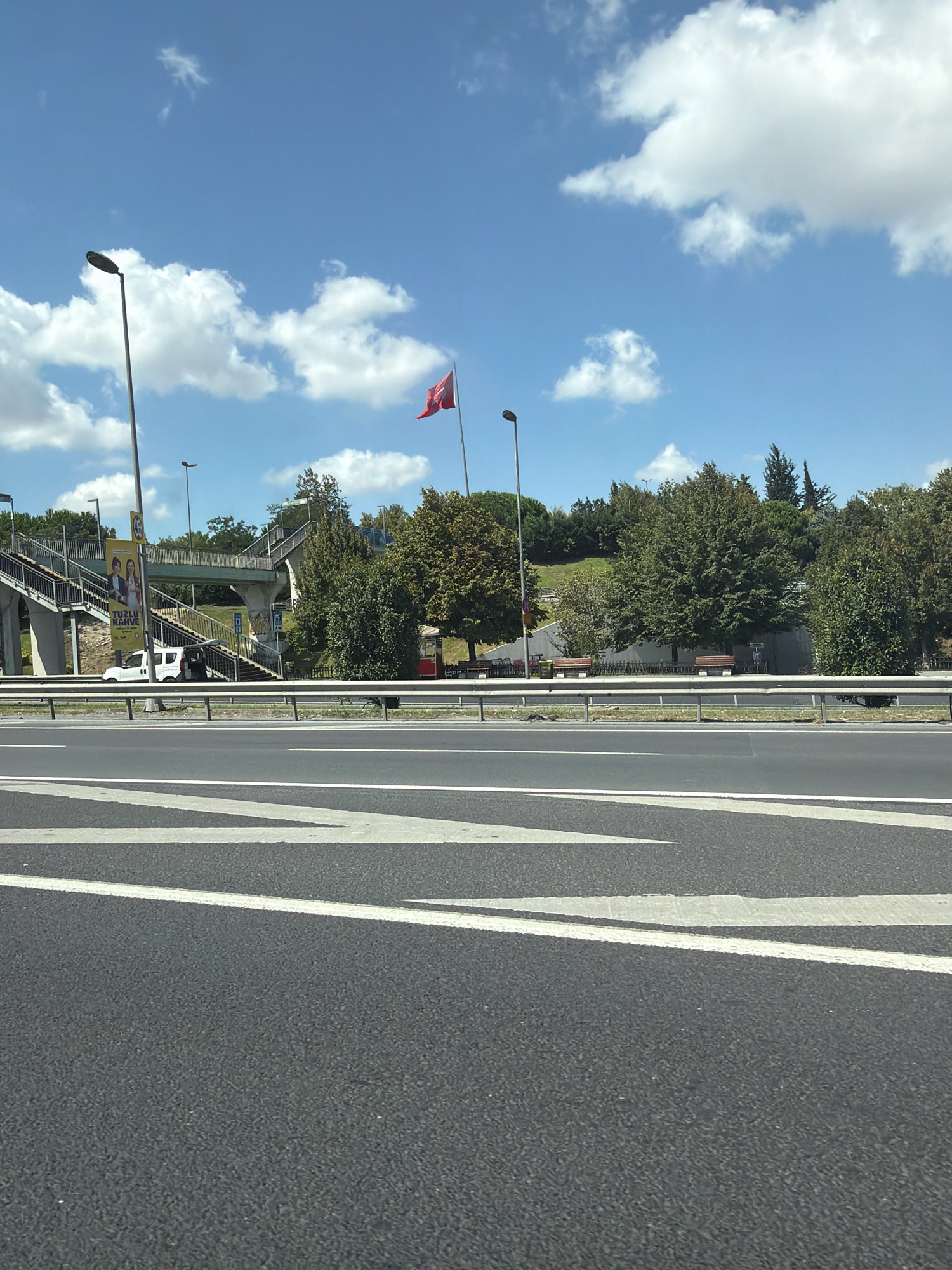 The image shows a bright, sunny day in Eyüpsultan, Turkey, with a clear blue sky dotted with fluffy white clouds. In the foreground, a section of a dark asphalt road with white lane markings is visible. Beyond the road, a metal guardrail separates it from a grassy embankment and a pedestrian overpass. The overpass, constructed of concrete and metal, has stairs leading up to it. A white van is parked near the base of the overpass, with its rear door open.
Further back, a Turkish flag is prominently displayed on a flagpole, waving in the breeze. The landscape is dotted with various trees, some with full green canopies and others appearing more sparse. A few park benches are visible near the overpass. A small red kiosk or structure is also present. A tall lamppost stands on the left side of the image. The overall scene depicts an urban environment with a focus on infrastructure and public spaces, under a clear, sunny sky.