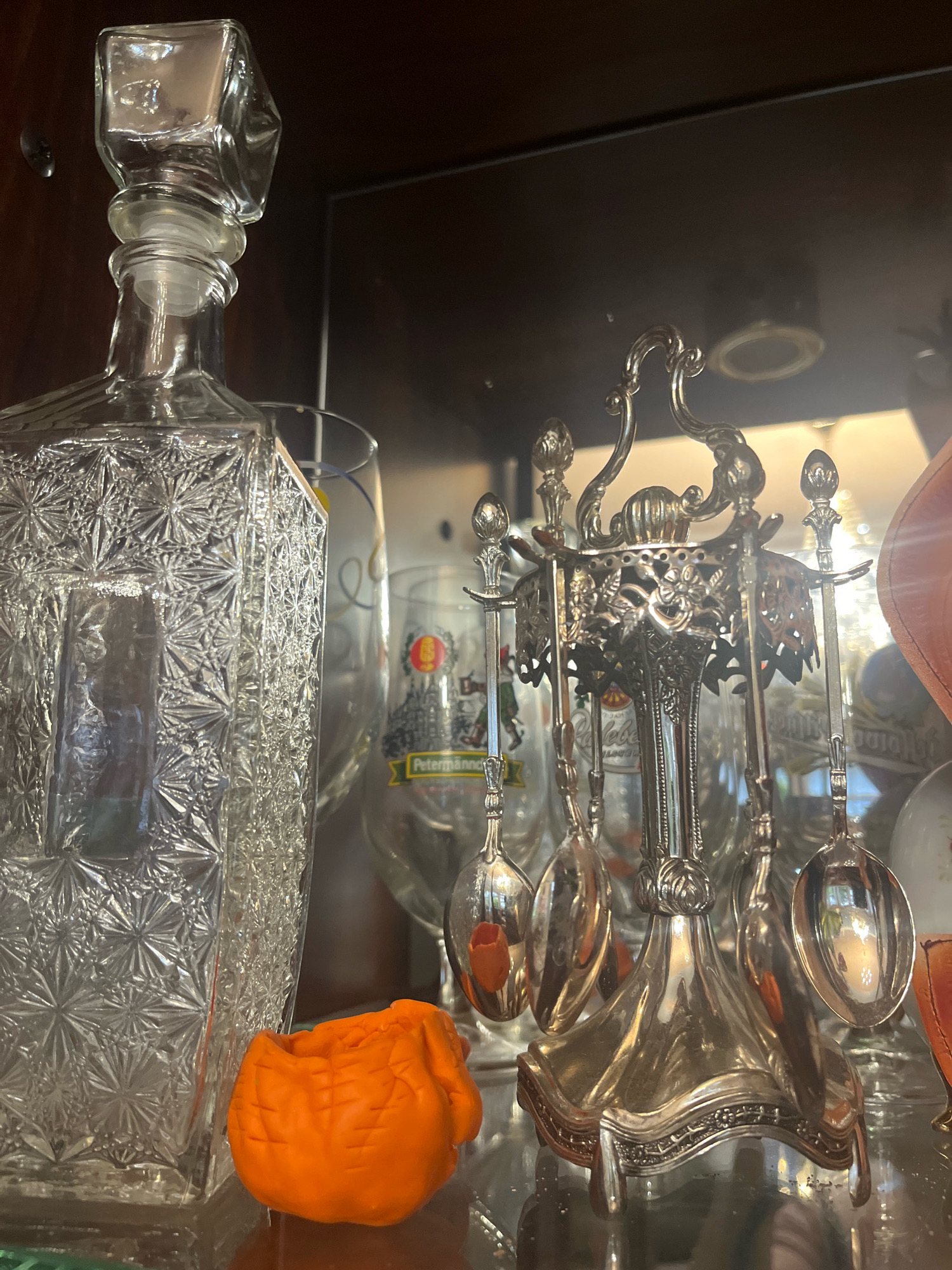A close-up, eye-level shot captures a collection of decorative items displayed on a glass shelf, possibly within a cabinet. Dominating the left side is a tall, clear glass decanter with an intricately patterned surface and a stopper. To the right of the decanter, a silver-plated spoon rack holds several small spoons. The rack features ornate scrollwork and floral motifs. In front of the spoon rack, partially visible, is a small, orange, carved object that resembles a pumpkin or a small fruit. In the background, several clear glass items, including what appear to be beer glasses with labels, are visible. The lighting suggests an indoor setting, possibly dimly lit with a warm glow from behind. The scene is devoid of any people or activity. The location is specified as Vladimirovka, Russia.