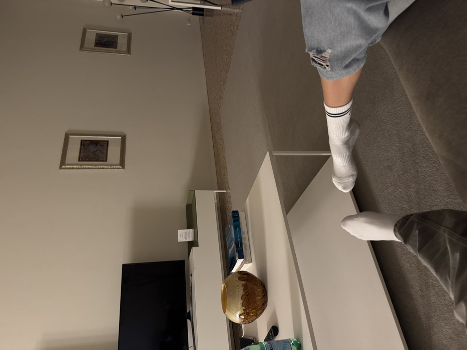 The image depicts an interior living space, identified as being in Venice, Italy, from a low-angle perspective, likely from a sofa. Two individuals are partially visible in the foreground with their legs extended. The person on the lower left wears light-colored pants and white socks. The person on the lower right wears ripped light blue jeans and white socks with black stripes, with their leg resting on a grey upholstered surface. A white rectangular coffee table with a lower shelf is centered in the mid-ground. On the coffee table are a dark amber-colored, spherical glass vase with a textured upper rim, two stacked books with blue and white spines, a green bottle (Pellegrino brand visible), and a black remote control.
Beyond the coffee table, a black flat-screen television is positioned on a white and light green media console against a light beige wall. A small white rectangular card is visible on the media console. Two framed artworks with ornate, gold-colored frames and dark central images are hung on the wall; one is above the console, and the other is further to the right. A modern, multi-branched black floor lamp with white globe-like lights stands in the far right background. The floor consists of a light grey carpet or rug covering the central area, surrounded by a light-colored speckled terrazzo-style hard floor. The room is uniformly lit, casting soft shadows.