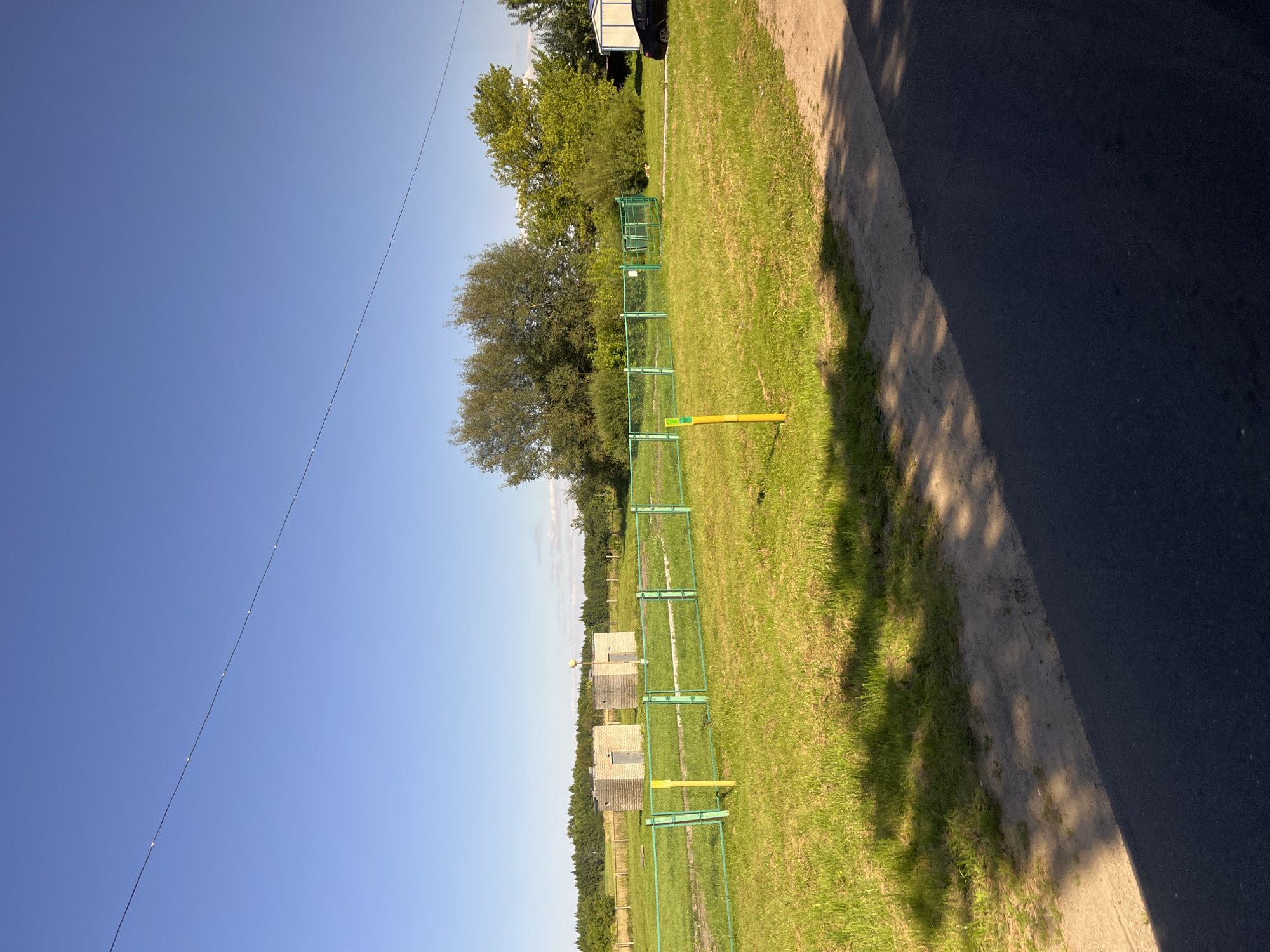 The image depicts an outdoor scene under a clear blue sky, likely on a sunny day. In the foreground, a paved surface, possibly a road or pathway, is visible, with a sandy or dirt shoulder. Lush green grass covers the majority of the mid-ground and background. A green metal fence with yellow posts delineates a section of the grassy area.
Within the fenced area, there are two small, rectangular brick structures with doors. Beyond the fence, a line of trees forms a natural boundary, with a dense forest visible in the distance. To the right of the fenced area, a white shed-like structure and a dark-colored car are present.
The scene suggests a rural or semi-rural setting, possibly on the outskirts of a settlement. The presence of the fence, the brick structures which could be utility buildings or storage, and the car indicate human activity. The overall impression is one of a quiet, possibly developing or established plot of land.
Based on the provided information, this scene is located in the city of Myadzyel, Belarus.