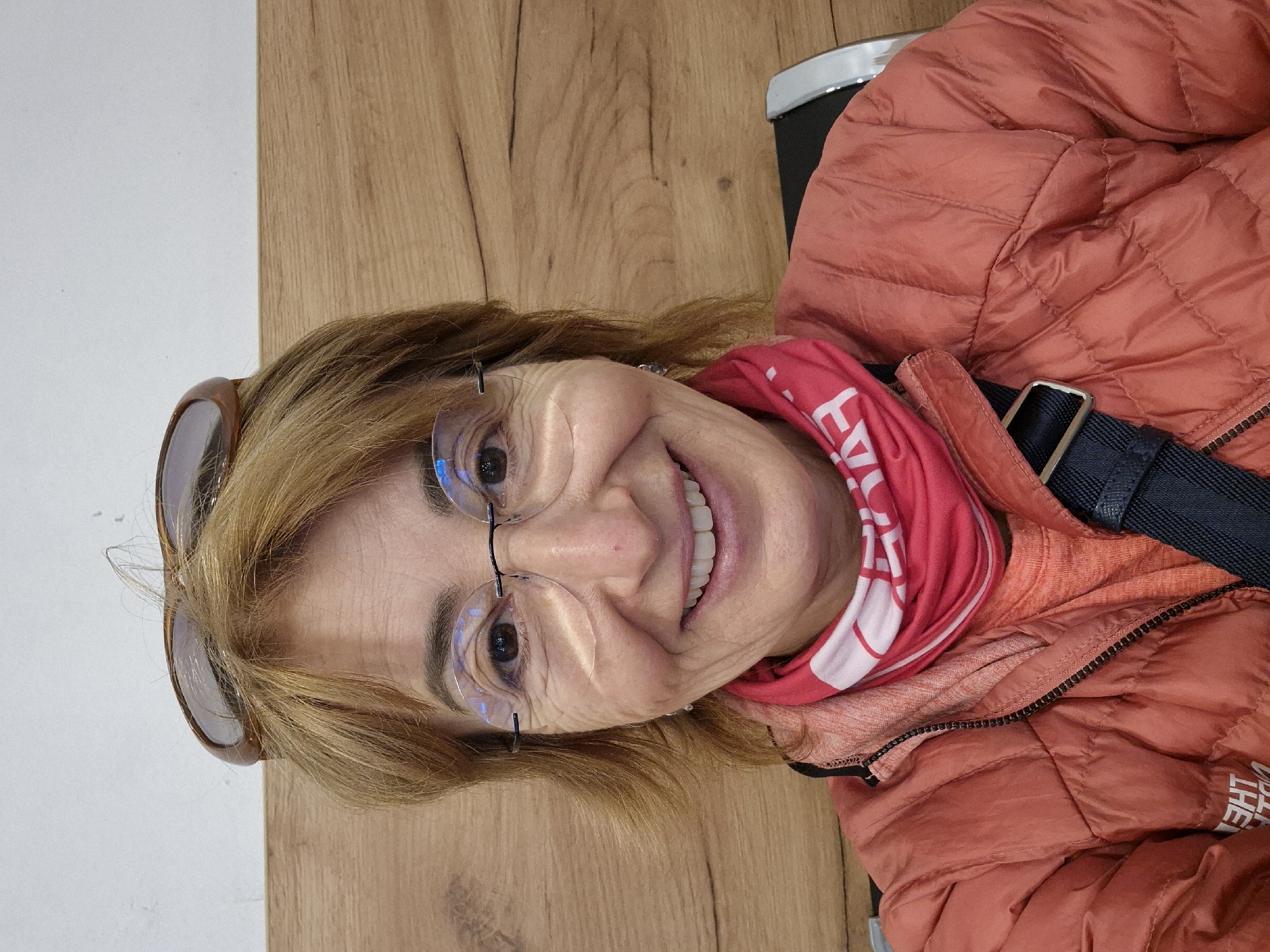 A cheerful, light-skinned woman is depicted in a close-up portrait, smiling broadly directly at the camera. She has light brown/blond, shoulder-length hair, with a pair of sunglasses resting on top of her head. She is wearing rimless eyeglasses. Her expression is happy and friendly.
She is dressed in warm, casual clothing, consisting of a reddish-orange puffy jacket and a red neck gaiter or buff around her neck. Visible text on the jacket, "THE", suggests a brand like "THE NORTH FACE". The neck gaiter also has white text, with "EV" partially visible. A black shoulder strap, likely from a bag, is visible across her chest.
The background is simple, divided between a light wooden surface on the right side of the frame and a plain white wall on the left, suggesting an indoor setting such as a cafe, waiting area, or similar public space. The warm attire implies a cooler climate. The overall scene captures a joyful moment, presumably in Vienna, Austria, as per the provided location context.