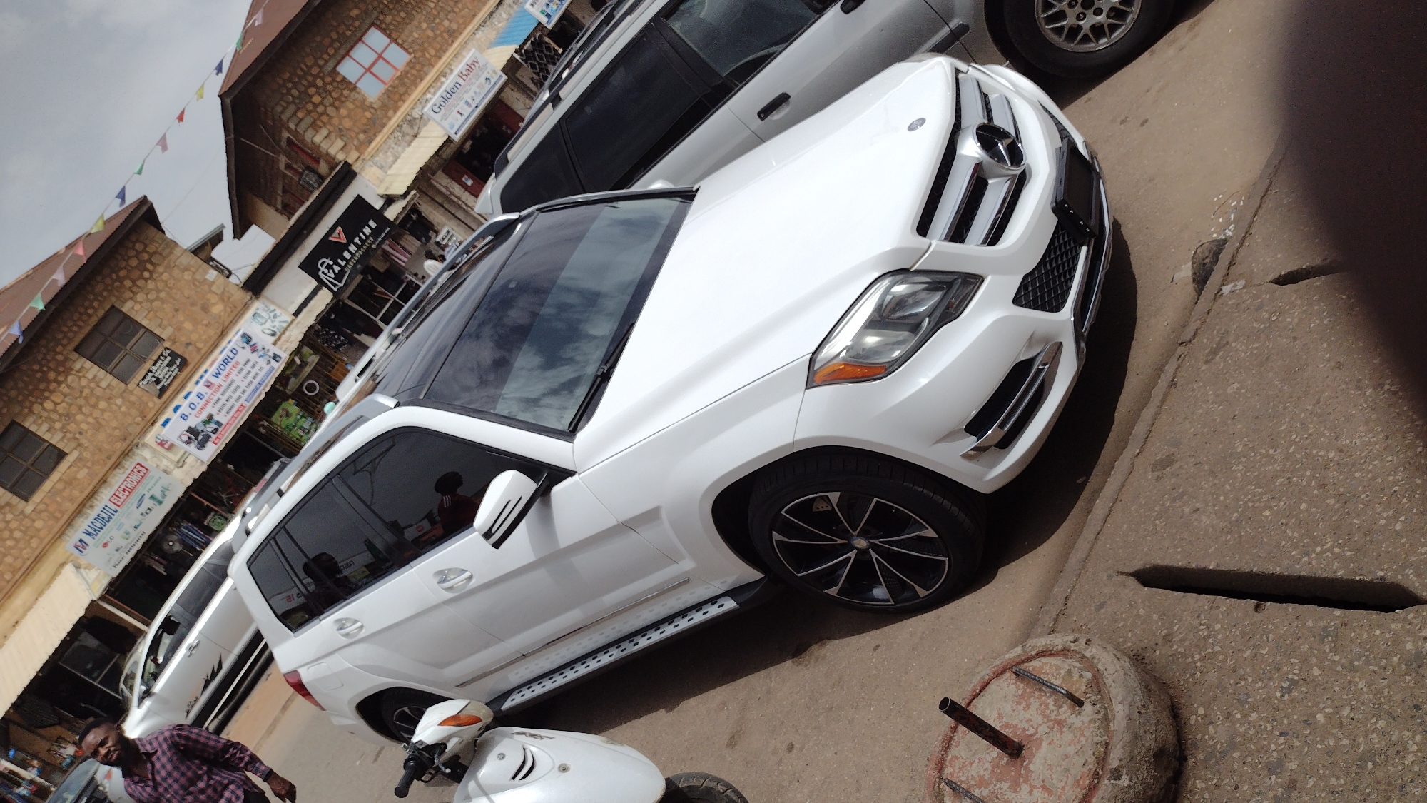 A white Mercedes-Benz SUV is parked on the side of a street lined with buildings featuring stone facades. The location is Jos, Nigeria, based on the presence of local businesses such as "B.O.B World Connection Limited" and "M. Macodji Electronics." A man in a maroon plaid shirt is walking on the sidewalk, partially visible in the lower left corner. A white moped or scooter is parked next to the SUV. A small, cylindrical concrete object with metal rods is on the sidewalk near the vehicle. The sky is overcast.