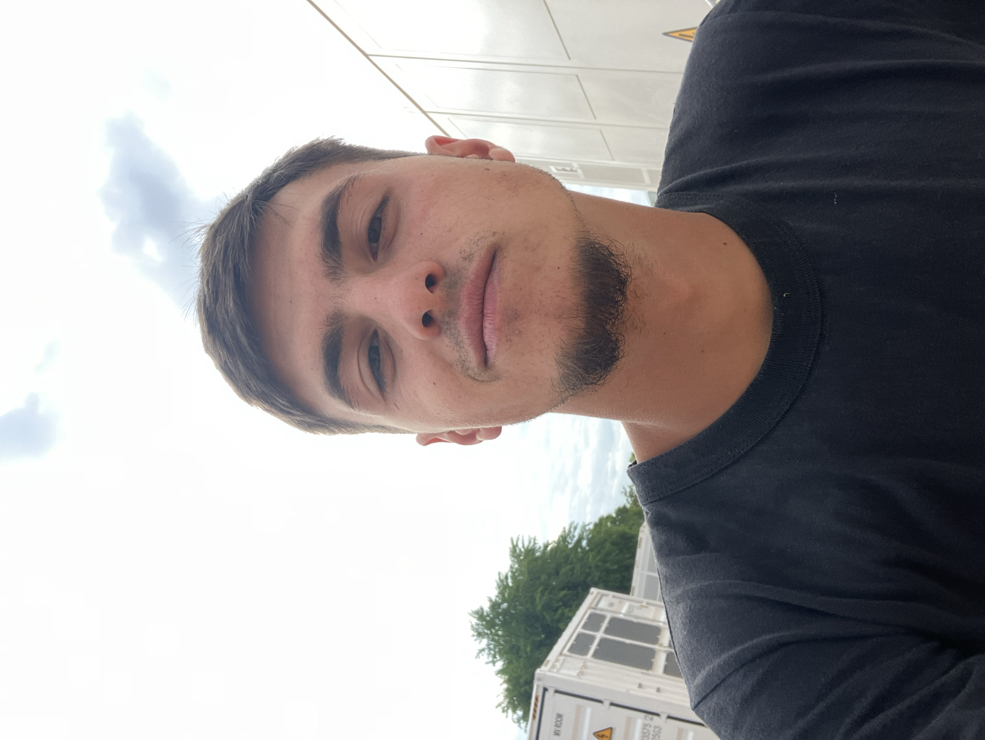 A man is shown in a close-up, selfie-style photo. He has short brown hair, fair skin, and a short, trimmed beard. He is wearing a black T-shirt. The background includes a bright sky with clouds, the top of a tree, and part of a white shipping container with warning labels and text (partially obscured). The location, Plovdiv, Bulgaria, cannot be verified from the provided image details.