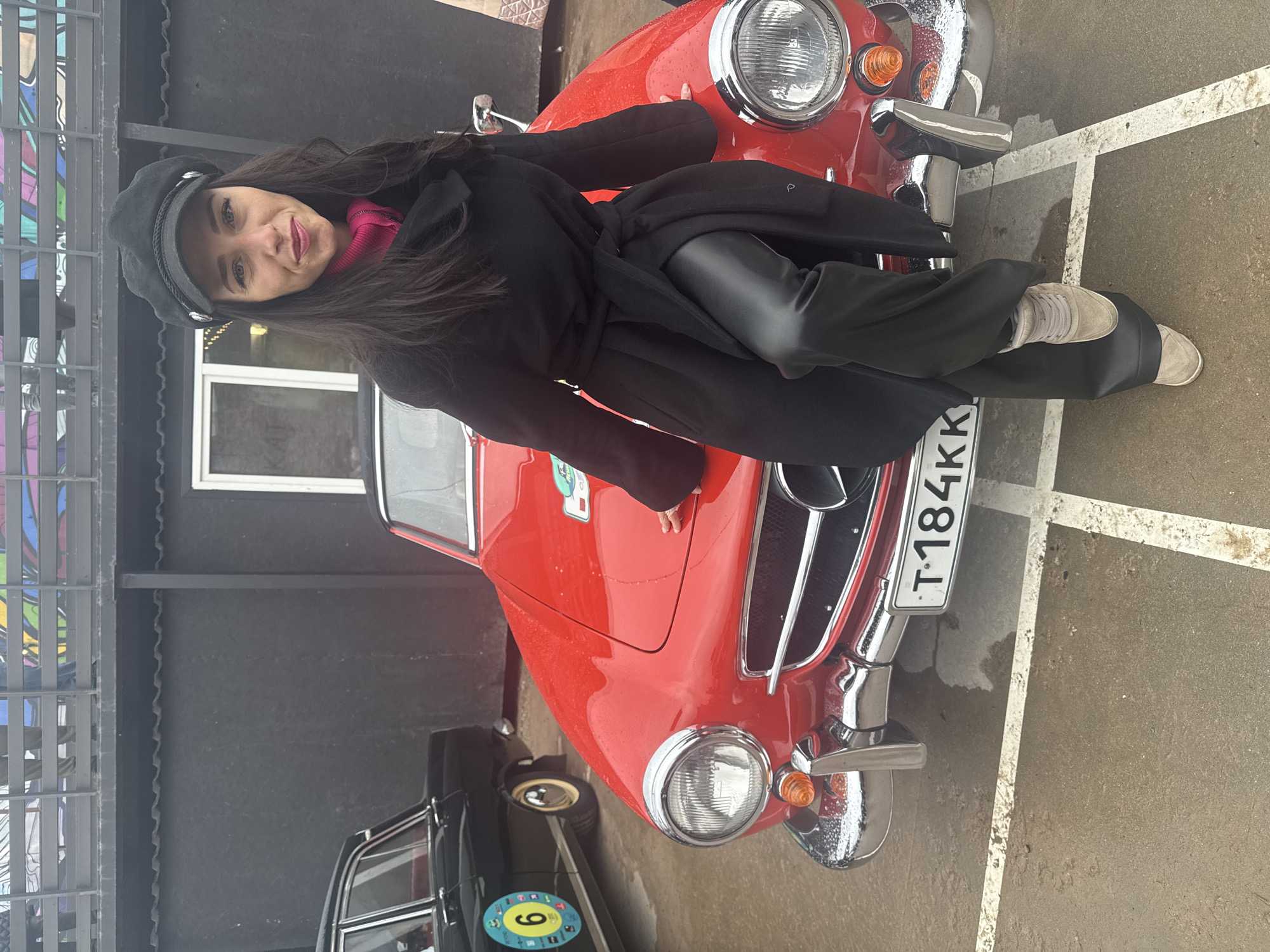 A woman in a black coat and hat is leaning on a red vintage car. The car's license plate reads "T184KK." Another, mostly black, car is partially visible behind the red one. The ground is paved. A building with a black facade and white window is in the background. The scene appears to be in Saint Petersburg, Russia.