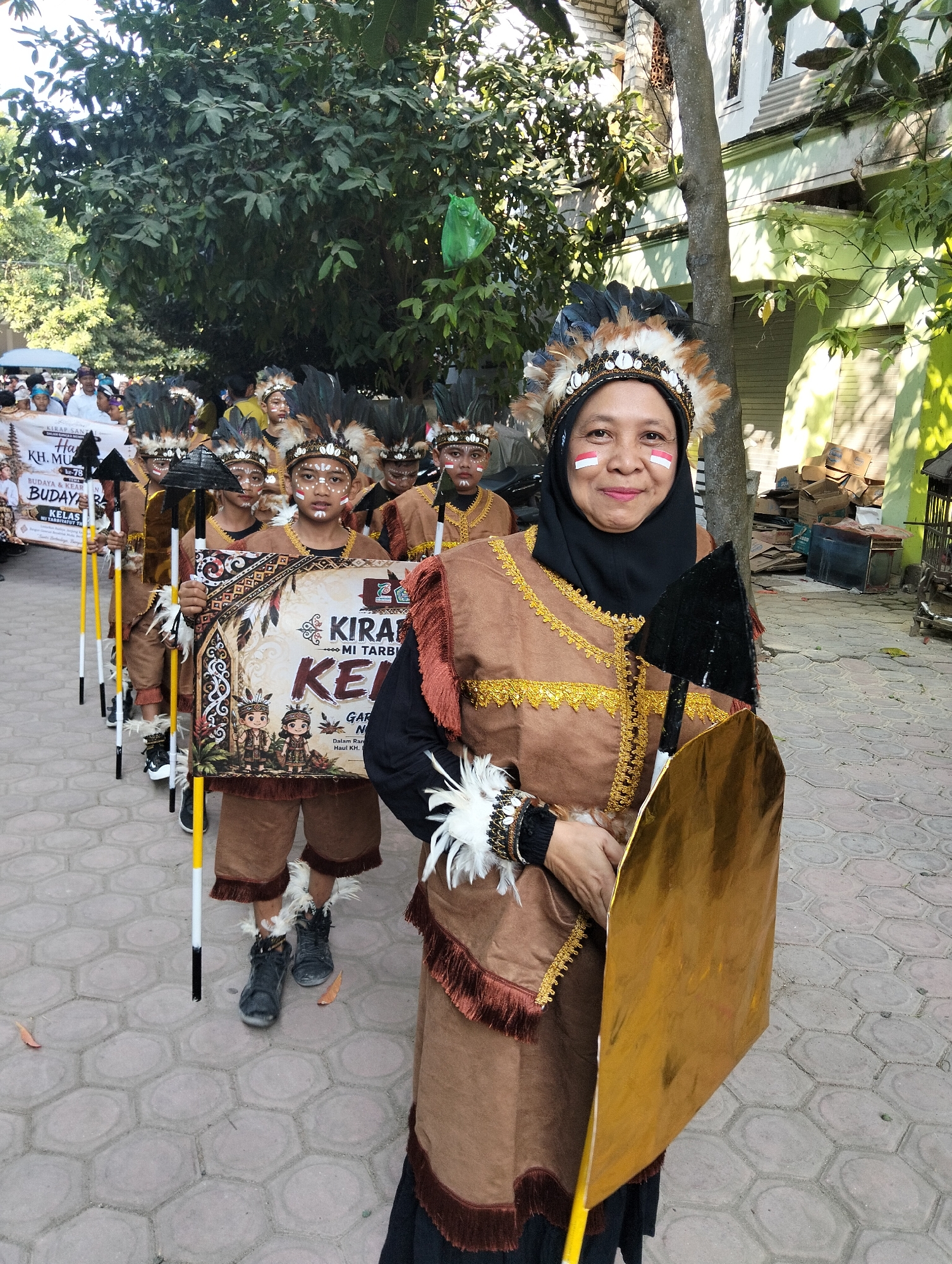 An outdoor procession is taking place on a brick walkway in Kranji, Indonesia. A woman in the foreground, wearing a black hijab, a brown tunic with gold trim, and a feathered headdress, holds a golden shield and a black spear. She has red and white face paint on her cheeks. Behind her, a group of children, also adorned in similar feathered headdresses and brown outfits, march in formation. Several children carry spears and one holds a banner. In the background, trees and buildings are visible, and a crowd of onlookers can be seen. The overall scene suggests a cultural event or parade.
