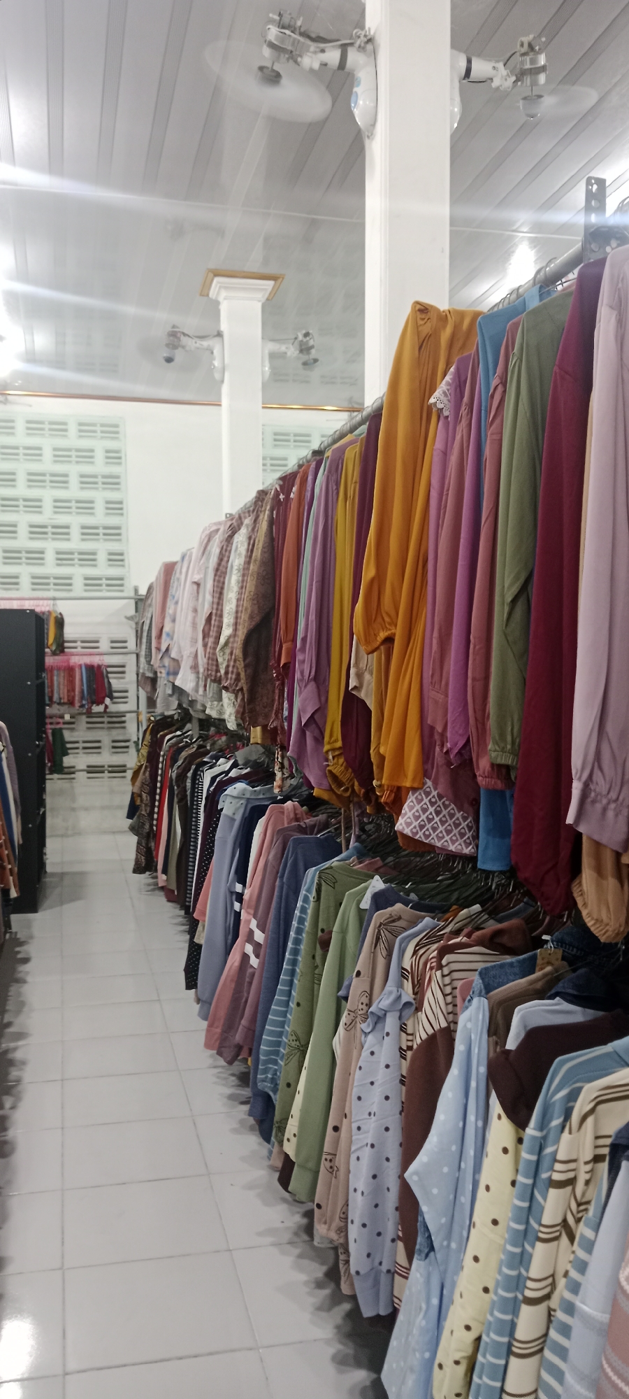 The image depicts a clothing store, likely in Rengel, Indonesia, with racks of apparel displayed on both sides of an aisle. The clothing, predominantly women's garments, hangs in a variety of colors and patterns. Some items are solid colored, while others feature stripes, polka dots, or floral motifs. The aisle is tiled with white square tiles. In the background, white wall vents and ceiling fans are visible, suggesting an indoor, possibly air-conditioned, retail environment. The lighting appears to be artificial, coming from overhead fluorescent fixtures. No people are visible in the image, focusing the attention entirely on the merchandise and the store's interior.