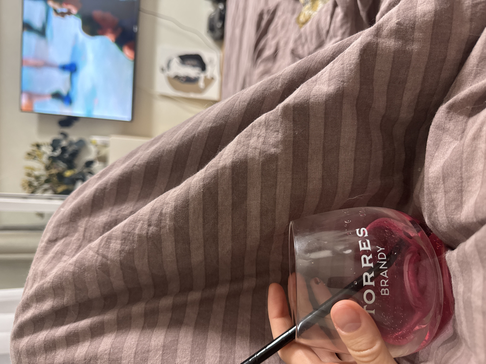 A low-angle, indoor shot features a hand holding a partially-filled "TORRES Brandy" glass with a black straw, resting on brown, striped bedding. A television displaying an indistinct scene, a decorative object, and a plant are visible in the blurred background. The location is suspected to be in the city of Chaikovsky, Russia.