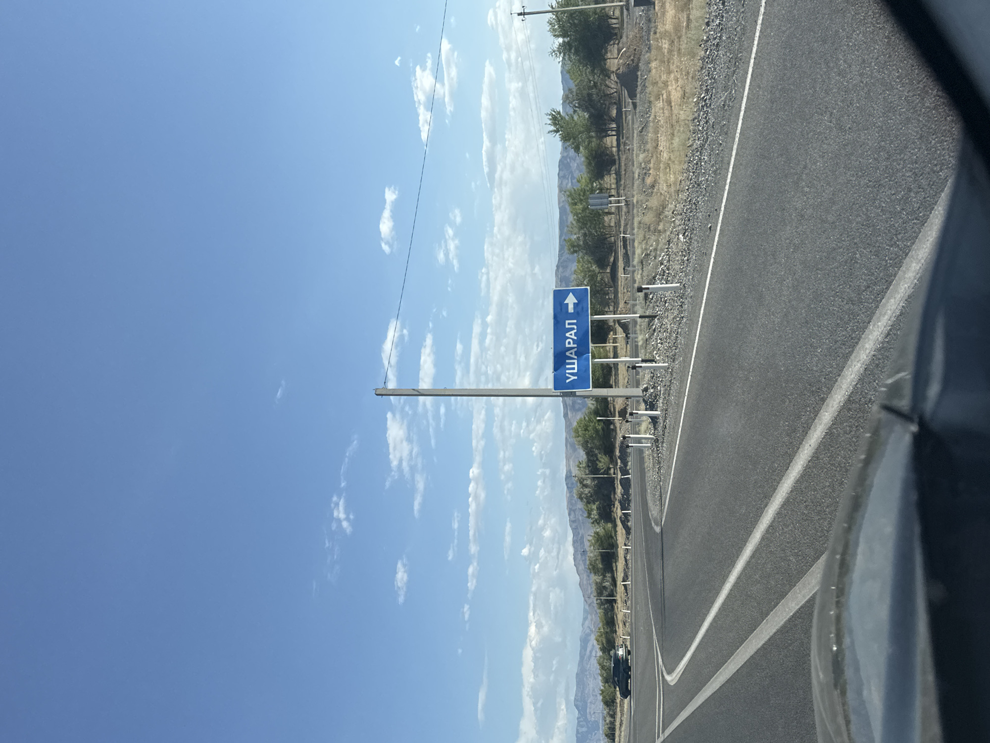 The image shows a road leading towards a highway intersection, with a blue sign indicating "Usharal" with a right-pointing arrow. The scene is set in a rural area under a clear blue sky with scattered clouds. In the foreground, the road surface is asphalt with white markings. A dark-colored vehicle is visible on the left lane of the road, turning. In the background, there are low, rolling hills covered in sparse vegetation and trees. A single utility pole stands next to the road, supporting a power line. The overall environment suggests a dry, possibly arid climate.