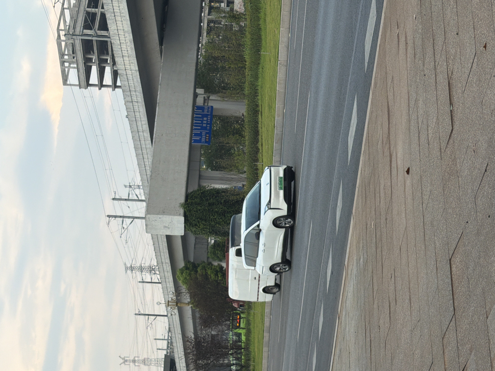 In the city of Jiangjin, China, a white minivan is seen driving on a paved road, likely during daylight hours. The minivan is in the right lane, with a white van in the left lane ahead of it. Behind the vehicles, a concrete overpass stretches across the scene, with a partially constructed building visible on the right side of the overpass. Power lines and towers are also present in the background, suggesting an urban or semi-urban environment with extensive infrastructure. The road has painted white lines indicating lanes and crosswalks. The surrounding area features a grassy median between the lanes and a paved pedestrian walkway on the left side of the road.
