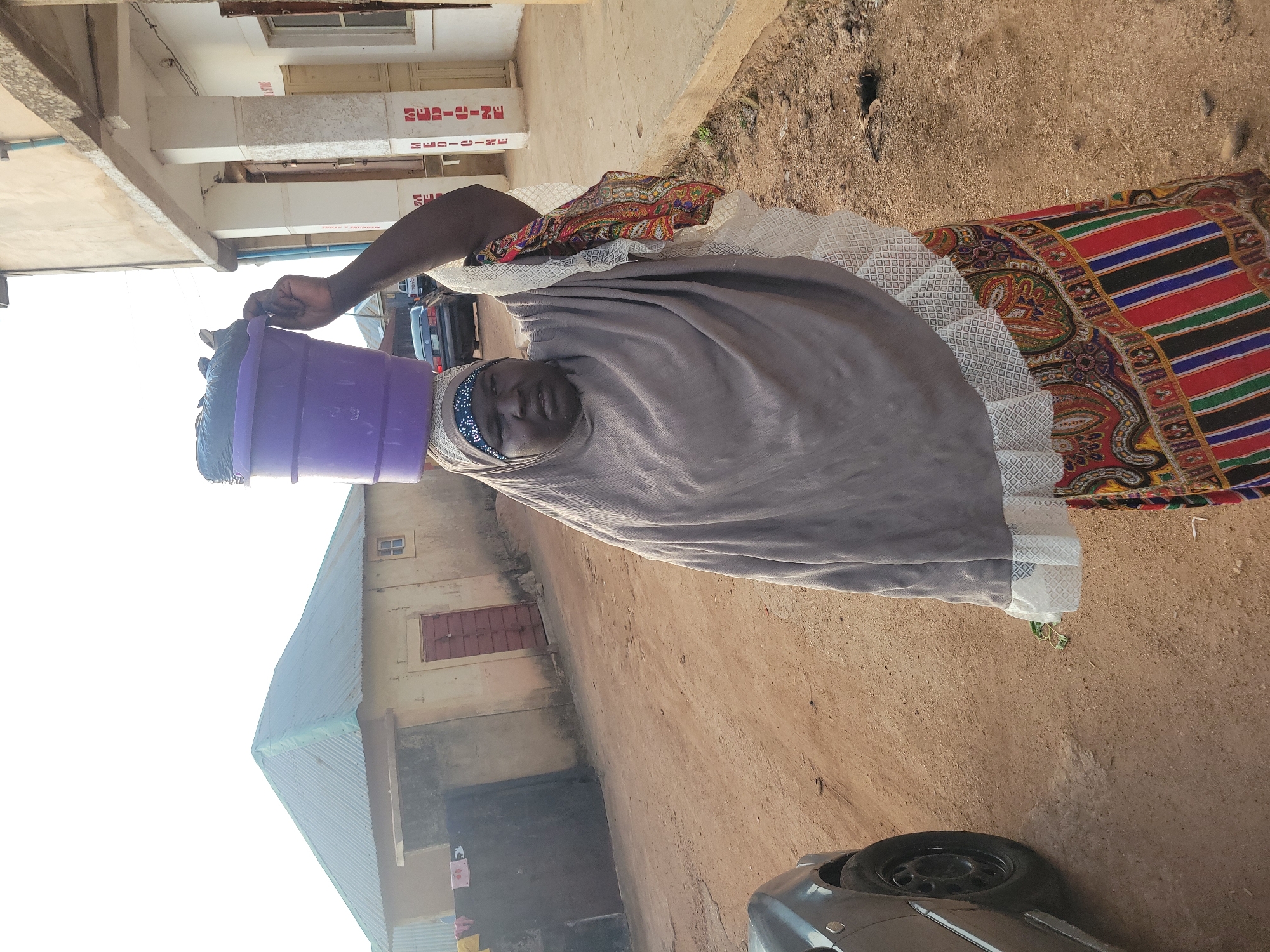 An adult woman stands on an unpaved dirt road in Jos, Nigeria, carrying a purple plastic bucket filled with dark material on her head, supported by her raised right hand. She wears a light grey hijab over a patterned inner headpiece and a colorful lower garment featuring red, blue, green, and black stripes along with paisley designs. A white, patterned trim is visible at the hem of her upper garment. To the immediate left, the front section of a dark-colored car is present. The road surface consists of reddish-brown earth with scattered debris. In the background, to the left, are two single-story buildings with light-colored walls and corrugated metal roofs; one features a dark gate, and the other a red door. Further down the road, a dark sedan and a white pickup truck are visible. To the right, a multi-story building with white concrete pillars stands adjacent to the road. The pillars bear vertical red text reading "MEDICINE" and "STORE". The sky above is bright and clear.