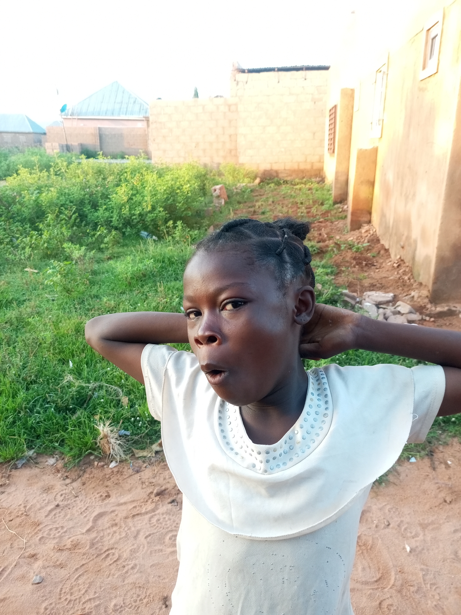 A young girl, appearing to be of African descent, with dark skin and braided hair pulled into two buns on top of her head, stands with her hands behind her neck. She is wearing a light-colored dress with a circular patterned neckline. Her mouth is open in a surprised expression. She is positioned in what appears to be an outdoor environment, with sandy ground and green vegetation in the background. Behind the vegetation, there are buildings constructed from concrete blocks, some with corrugated metal roofs. One building has a visible satellite dish. The overall impression is a scene in a semi-urban or rural setting in Tempure, Nigeria. The lighting suggests it is daytime, possibly late afternoon due to the warm tone of the light.