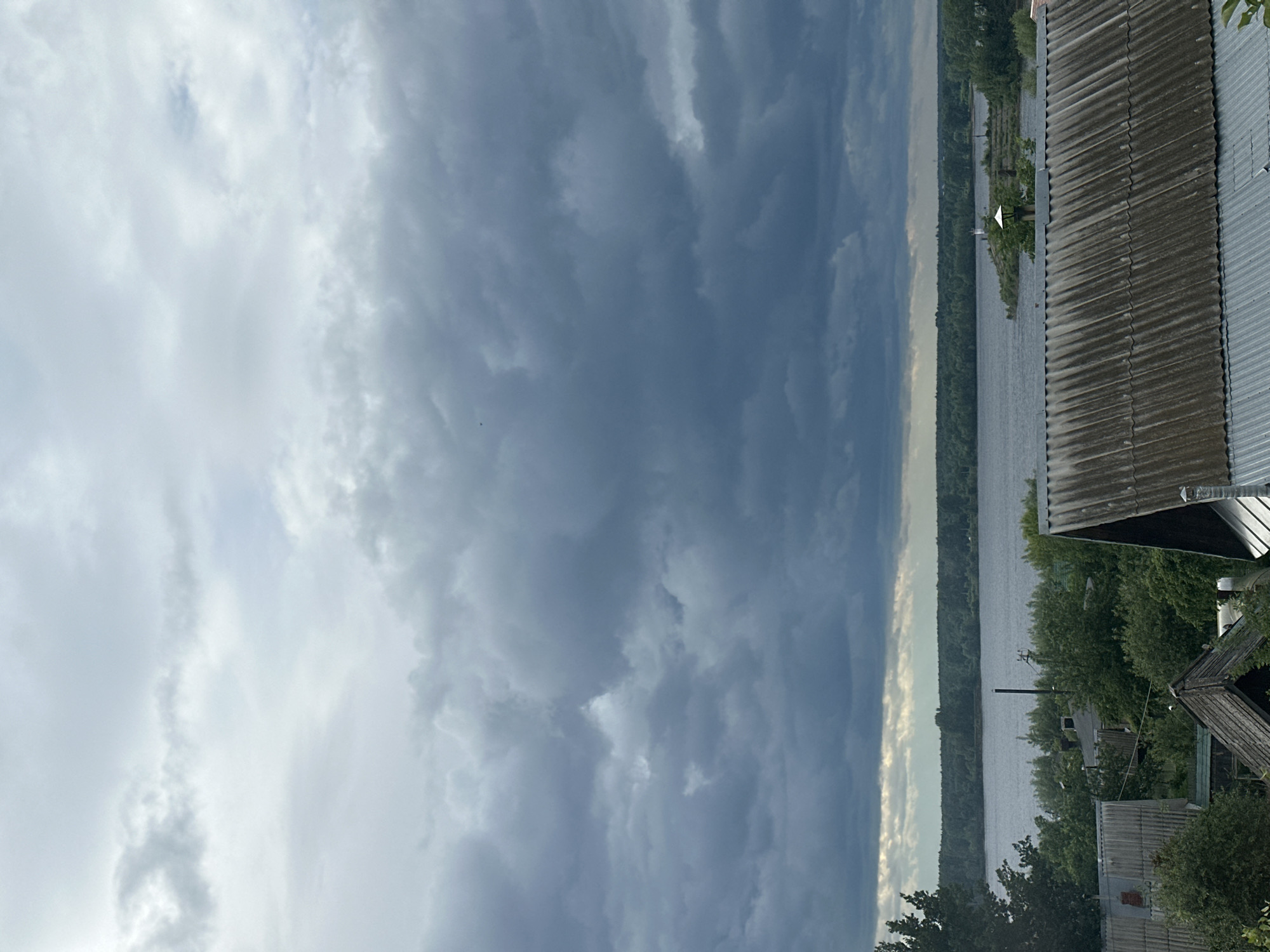 This landscape image, likely taken in Chaikovsky, Russia, captures a wide expanse of water beneath a dynamic, cloud-filled sky. The scene is oriented horizontally (when viewed correctly), with the sky dominating the upper half. Dramatic clouds range from bright, soft white on the left to darker, more voluminous grey and blue formations on the right, suggesting varying weather conditions or an approaching front.
Below the sky, a wide river or lake stretches across the middle of the frame, its surface reflecting the overhead light. The far bank is lined with a dense, dark green forest that appears to be vast and continuous.
On the near bank, visible on the right, several rustic buildings are nestled among trees. These structures feature corrugated metal or slate roofs, some appearing weathered. Utility poles are also faintly visible on this side of the bank.
The time of day appears to be daytime, possibly late afternoon, characterized by the strong contrast in the clouds and the reflective quality of the water. The overall impression is one of serene natural beauty with a hint of dramatic weather. No people are visible, and there is no discernible activity or text within the image.