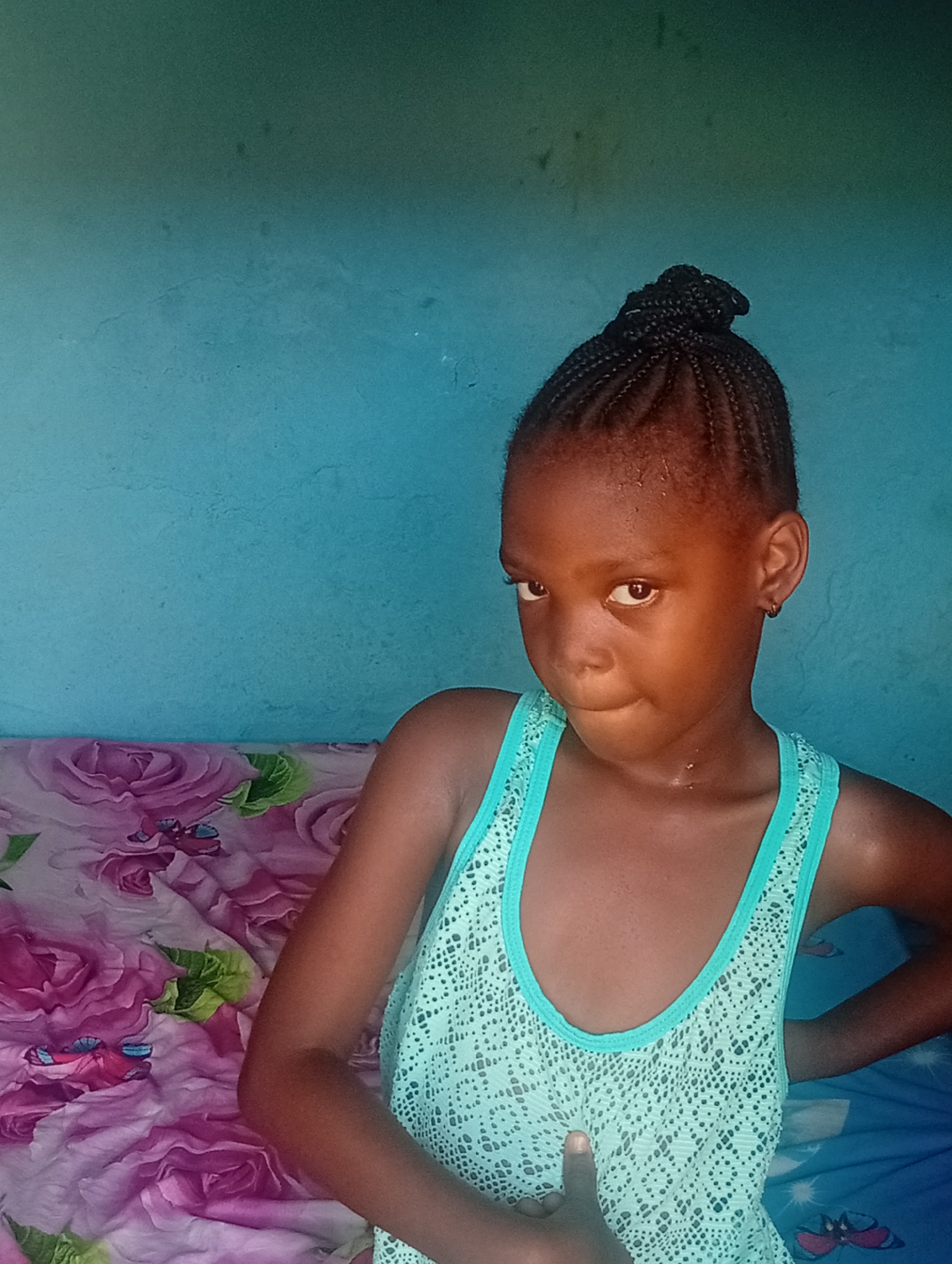 A young girl with braided hair pulled into a bun stands indoors. She wears a light blue, patterned tank top. Her expression is somewhat quizzical or serious. She has one hand on her hip. Behind her is a blue wall and a bed with a floral pattern. The location is Unknown, Unknown.