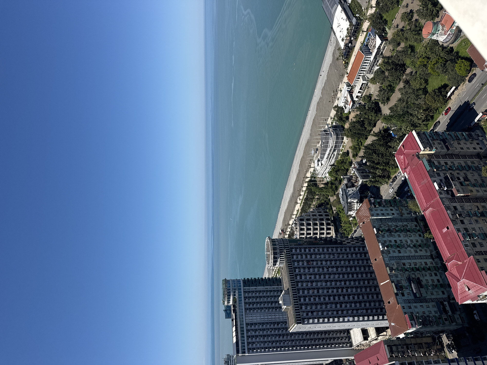 The image shows an aerial view of Batumi, Georgia. The Black Sea stretches along the left side of the frame, with a beach adjacent to the water. Beyond the beach, a line of trees and buildings runs along the coastline. In the lower-right corner of the image are several tall buildings with red roofs. The sky above the sea is clear and blue.