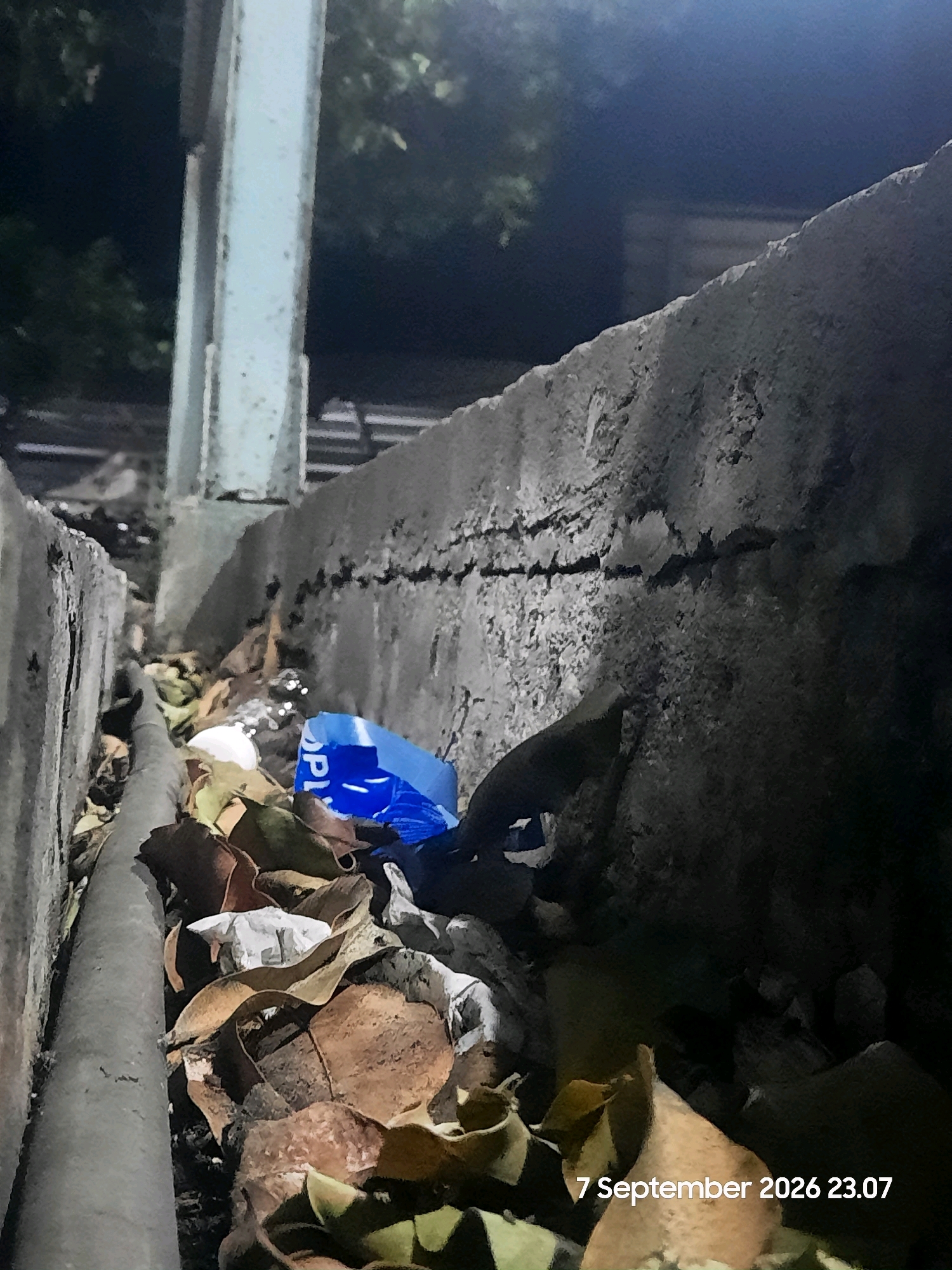 The image depicts an outdoor scene at night, possibly a drainage ditch or a narrow alleyway. A large, grey concrete structure with visible cracks runs diagonally across the frame, forming a wall. To the left, a vertical, pale blue metal beam is visible. The ground is covered with debris, including dry leaves, crumpled white paper, and a bright blue plastic bag with some lettering. A dark, cylindrical object, possibly a pipe or hose, runs along the bottom left of the image. The lighting suggests artificial illumination from above and to the right, casting shadows and highlighting the textures of the concrete and debris. The timestamp "7 September 2026 23.07" is superimposed in the bottom right corner. There are no people or animals visible in the image. The environment appears unkempt and possibly neglected.