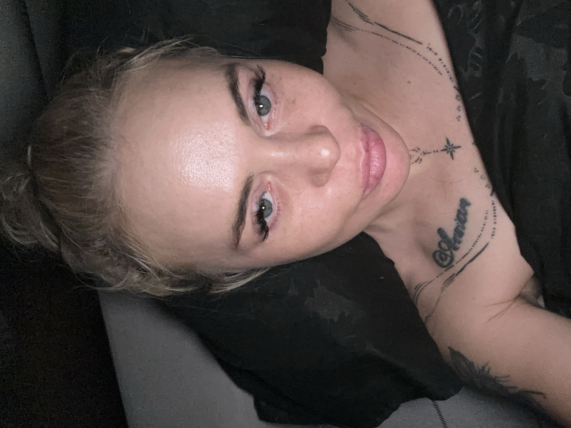 A close-up, eye-level portrait captures an adult female from the chest up, positioned in a dimly lit indoor setting at an unknown location in Unknown, Unknown. The subject, with light skin and blonde hair tied in a messy bun, looks directly into the camera. Her light blue eyes are prominent, enhanced by long, dark false eyelashes. Her lips are full, reddish-pink, and pursed into a slight pout. Visible skin shows scattered freckles or minor discolorations, with some sheen on the forehead.
Multiple tattoos are visible on her upper torso: on her right collarbone and upper chest, the script word "@Anna" is present, underlined by a series of small dots and linear designs. Below her neck, centrally located, is a small, stylized star connected by a linear element above it. A dark, possibly abstract or floral tattoo is partially visible on her left upper arm/shoulder. The woman's shoulders appear bare, and she is covered by or lying against dark, indistinct fabric, likely bedding. The background is uniformly dark, suggesting low light conditions with illumination coming from the front, casting soft shadows.