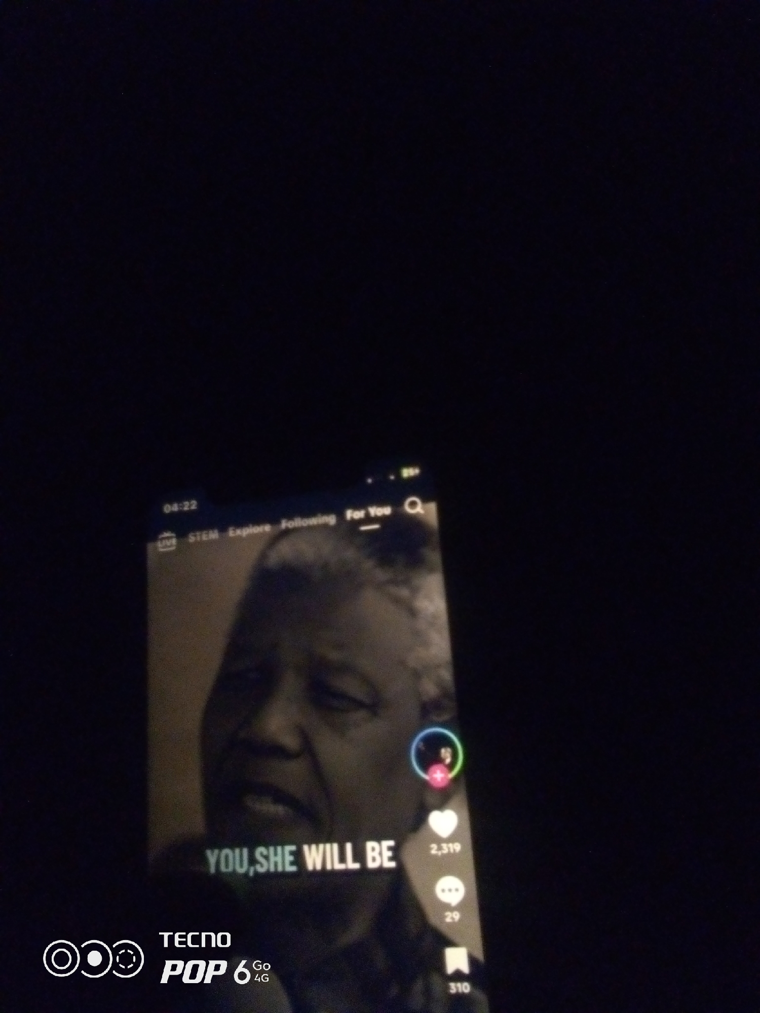 The image is a poorly lit photograph of a smartphone screen displaying a video. The video appears to be a clip of Nelson Mandela speaking, with superimposed text that reads "YOU, SHE WILL BE". On the bottom left of the screen, there is a logo that says "TECNO POP 6 Go 4G". The phone screen is slightly angled and the lighting is very dim, suggesting the photo was taken in a dark environment. There are no specific details in the image that can indicate the location in the city of Howayi, Nigeria.