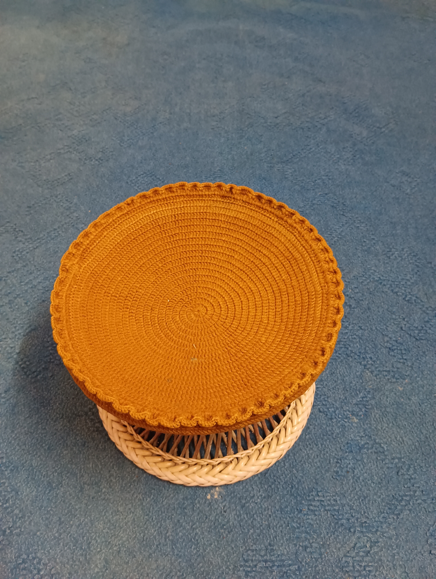 A close-up, overhead shot shows a round, woven orange placemat with a scalloped edge resting on a white woven base. The placemat is intricately textured with concentric circles of stitches, creating a spiral pattern that emanates from the center. The white base is also woven, with a criss-cross pattern visible through its open structure. Both items are placed on a textured blue surface, possibly a rug or fabric, which fills the background. The lighting is soft, casting a gentle shadow from the placemat onto the blue surface. There are no people or any indication of a specific location within Manali, India.
