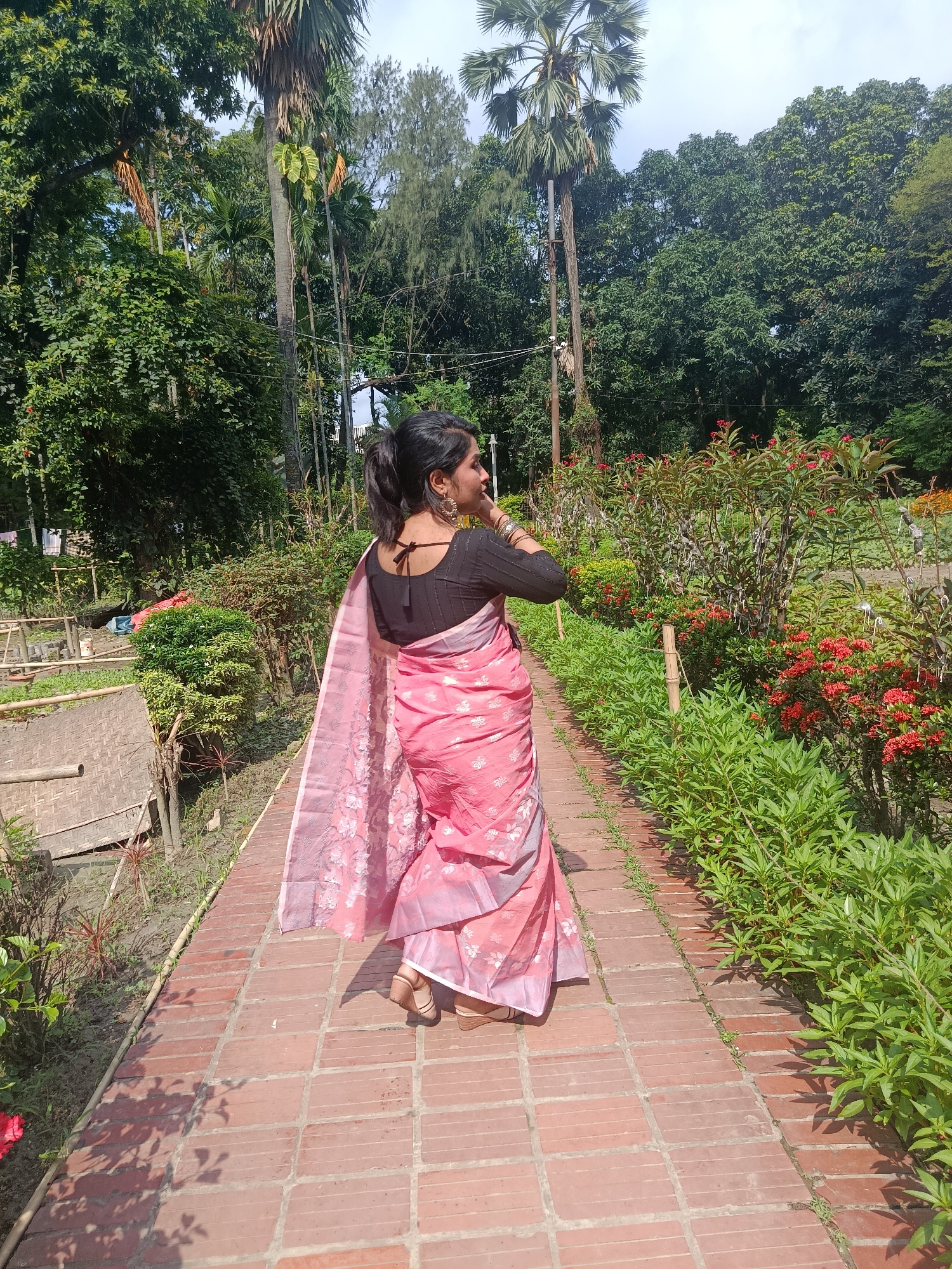 A woman is standing on a brick path in a garden. She is wearing a pink sari with a black blouse, her hair is up in a ponytail. She is facing away from the camera, looking over her right shoulder. There are green bushes and red flowers on either side of the path. Behind her are trees and foliage. The photo was taken in the city of Dhaka, Bangladesh.