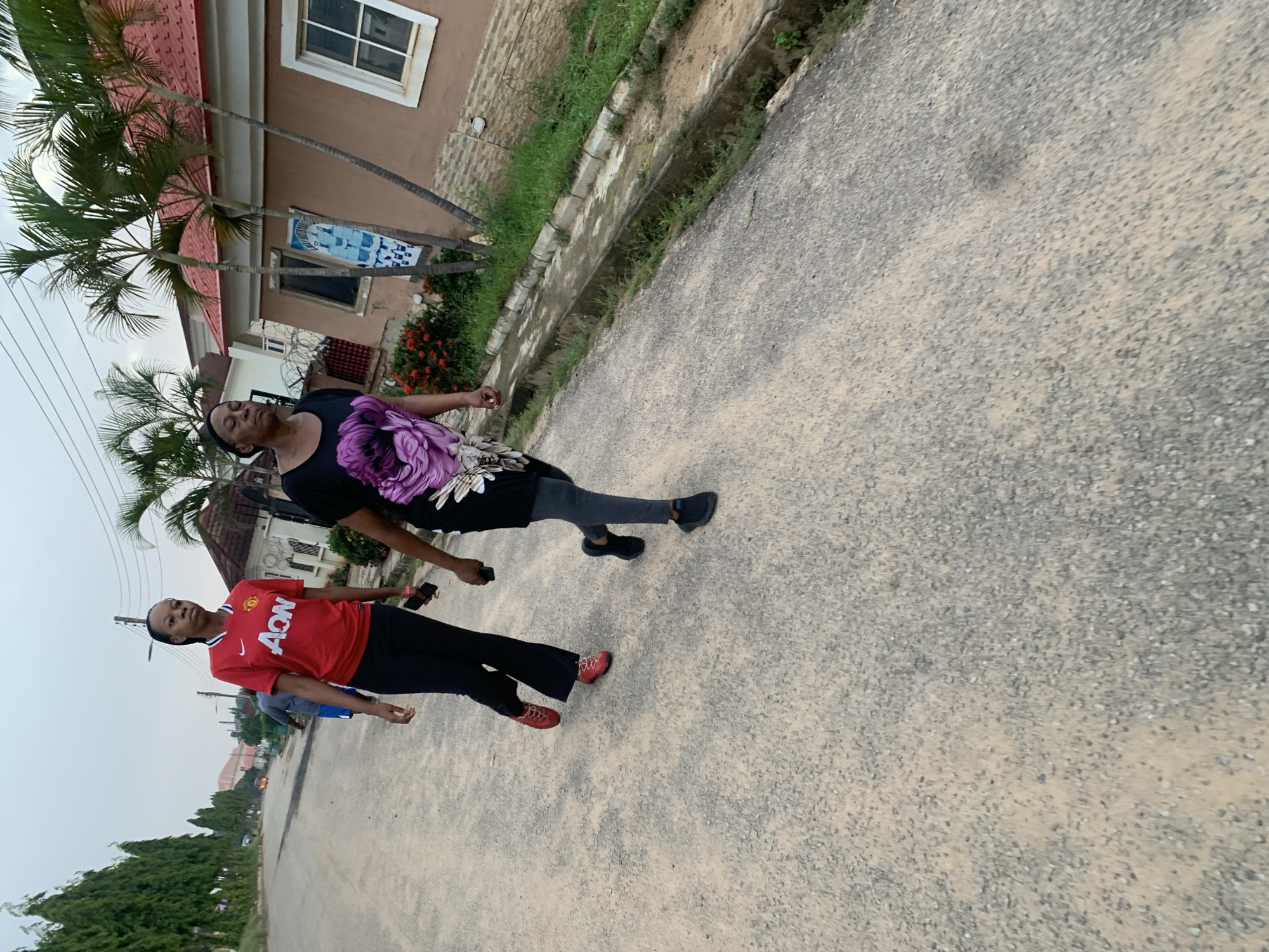 Two women walk on a dusty, asphalt road in Abuja, Nigeria. The woman on the left wears a red Manchester United jersey and black pants. The woman on the right wears a black t-shirt with a large purple floral design and dark leggings. They are walking away from the camera, towards residential buildings with red roofs. Palm trees and lush green vegetation surround the houses. The sky is overcast.