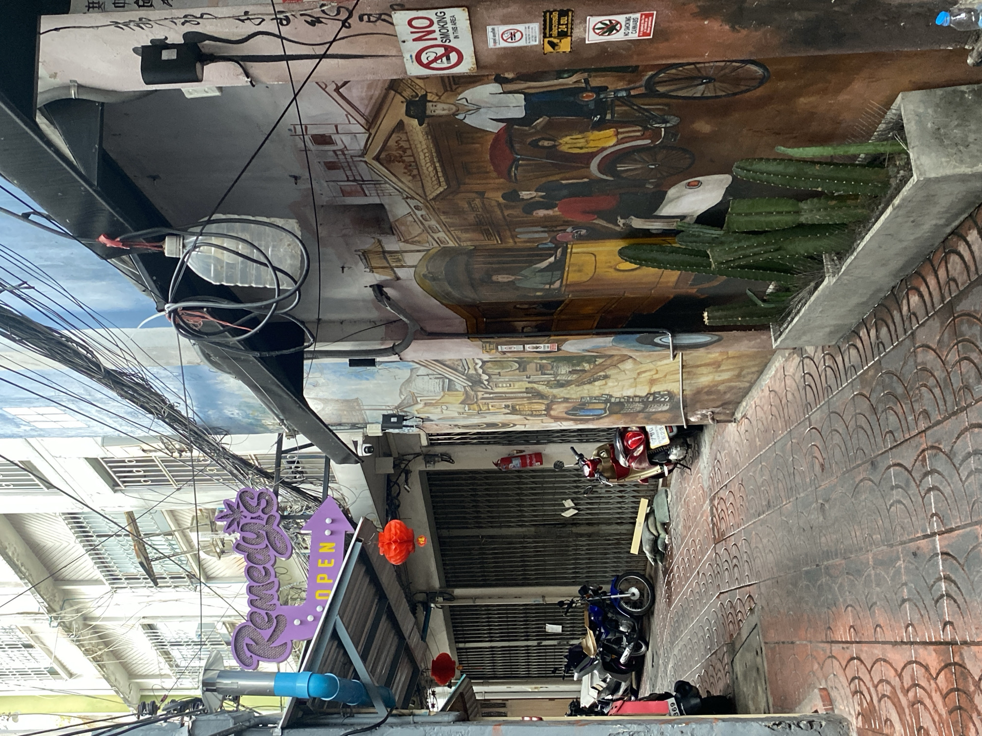This outdoor shot depicts a narrow alley in Bangkok, Thailand. The image is oriented vertically. The walls of the alley are adorned with murals depicting scenes of Bangkok, including historical figures on bicycles and in rickshaws. There are signs prohibiting smoking and cannabis use. The alley floor is paved with decorated tiles. Parked motorcycles occupy the space between the buildings on either side of the alley. Wires crisscross the upper part of the frame. A purple sign with "Remedy's" and an arrow pointing to "Open" is visible on the left.