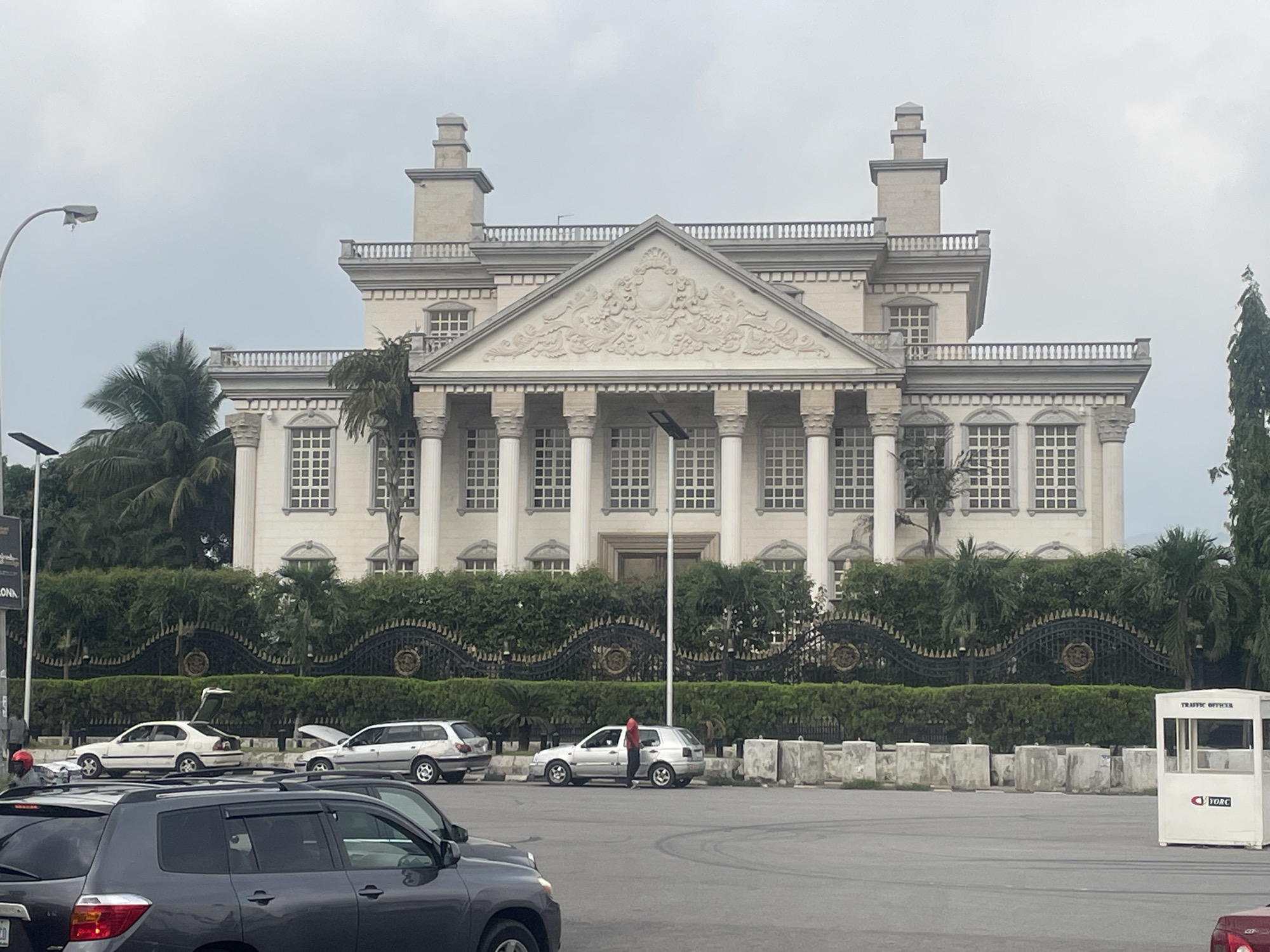 In Abuja, Nigeria, a grand, neoclassical mansion stands prominently under an overcast sky. The building features a symmetrical facade with a central triangular pediment adorned with intricate relief carvings. Rows of tall, white columns support the upper levels, and numerous square-paned windows are evenly spaced across the exterior.
In front of the mansion, a thick, well-maintained hedge runs the length of the property, bordered by an ornate black metal fence with decorative circular motifs. Several vehicles are parked or stopped on the paved area before the hedge. Among them is a silver hatchback with its hood open, and a white sedan with its trunk ajar. A person in a red shirt is standing next to the silver hatchback. Further to the right, a small white booth labeled "TRAFFIC OFFICER" with a logo is visible.
The foreground of the image shows the front of several dark-colored SUVs, suggesting a busy street or parking area. To the left, a lamppost with a solar panel is positioned. Trees, including palm trees and a tall evergreen, are scattered around the property, some growing near the mansion's facade and others taller in the background. The overall impression is one of opulence and security.