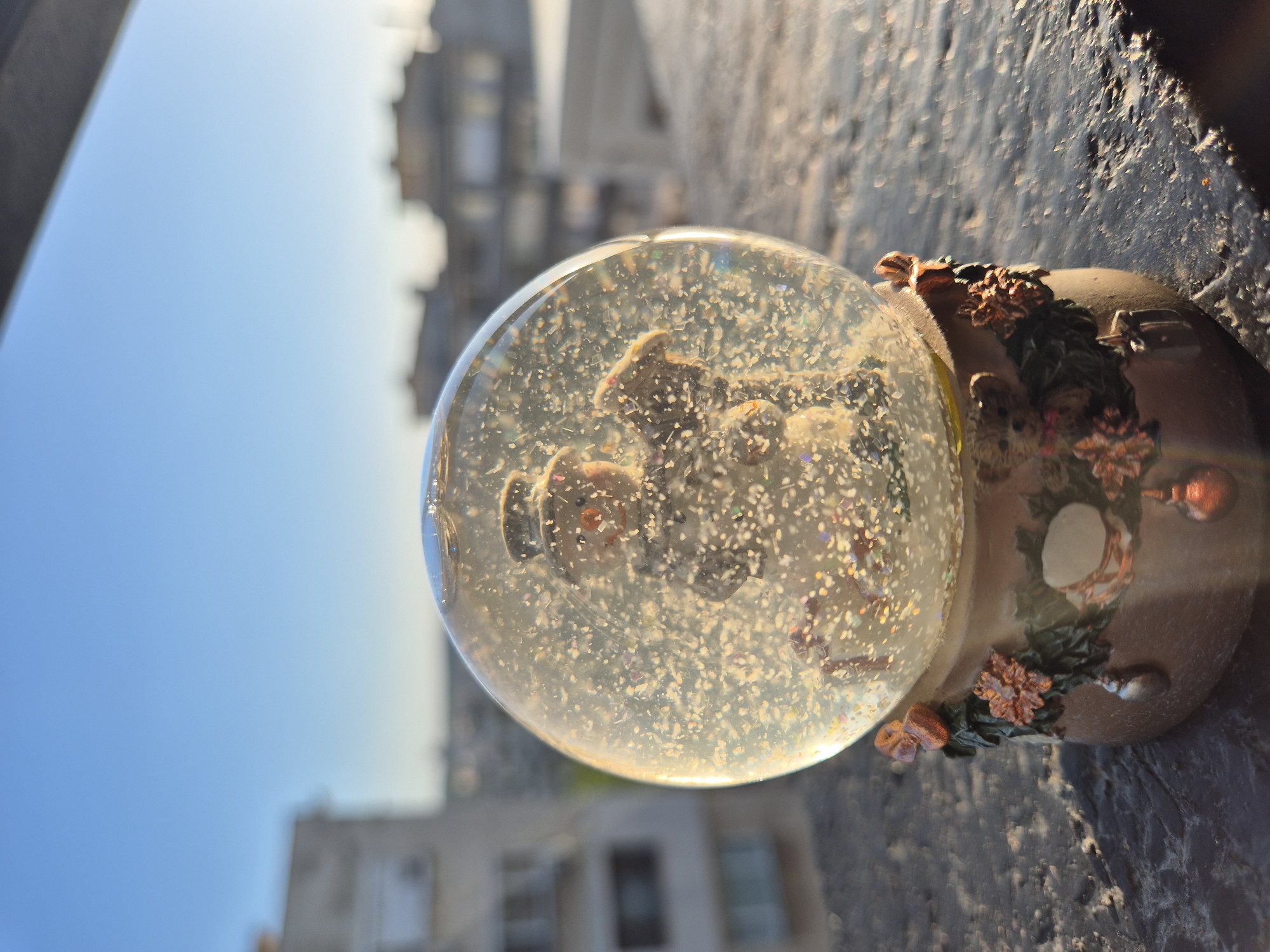 A snow globe sits on a concrete surface, with a blurred cityscape in the background. The snow globe's base is decorated with flowers and leaves, while the globe itself contains figures and glitter. Sunlight illuminates the scene, casting shadows and creating a warm glow. The location appears to be outdoors, possibly on a ledge or balcony in an urban setting, city of Unknown, Unknown.
