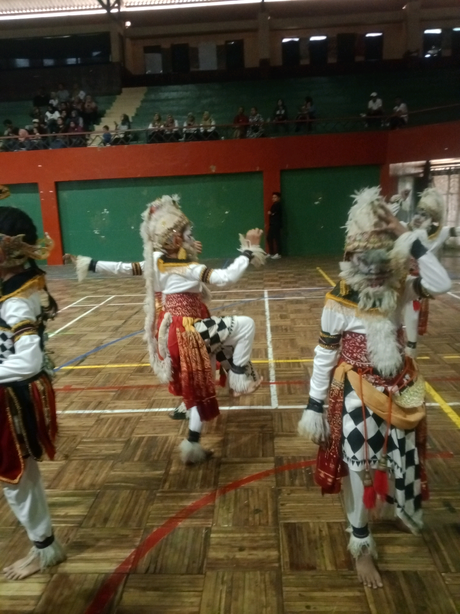 The image depicts a cultural performance taking place on a wooden gymnasium floor in Wuryorejo, Indonesia. Three individuals, adorned in elaborate traditional costumes, are the primary subjects. Their attire features white tops with gold trim, red and patterned skirts, and distinctive headdresses resembling animal fur with golden embellishments. The costumes also include white pants, black and white checkered patterns, and furry accents around the ankles and wrists, with some individuals appearing barefoot.
The performers are captured mid-motion, suggesting an active dance routine. The background reveals a tiered seating area, where spectators are visible, some seated and others standing. The seating area is characterized by green and orange sections. The gymnasium setting is further indicated by the presence of court lines on the floor. A person in dark clothing is standing in the background to the left of the central performers, observing the event. The overall lighting suggests an indoor environment, possibly under artificial lights.