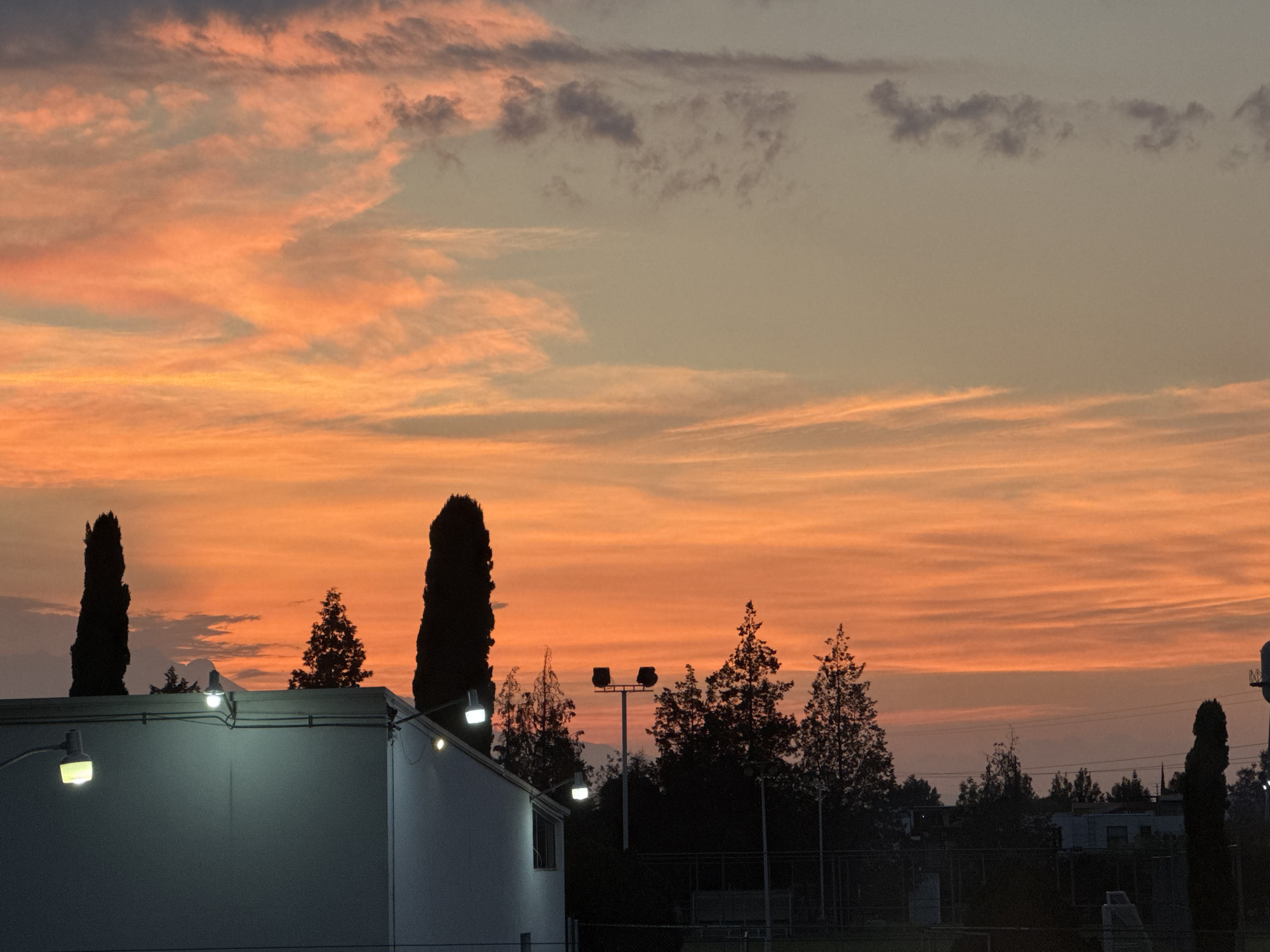 The image shows a dusk scene in Heroica Puebla de Zaragoza, Mexico. The sky is a mix of orange and gray, with streaky clouds. The foreground features a one-story building with exterior lights illuminated. Behind the building are dark silhouettes of trees and possibly light poles. Two tall, thin trees are prominent on the left and right sides of the frame. The rest of the scene includes houses in the background. A fence is visible in the lower part of the photo. The time of day appears to be either late evening or early morning.