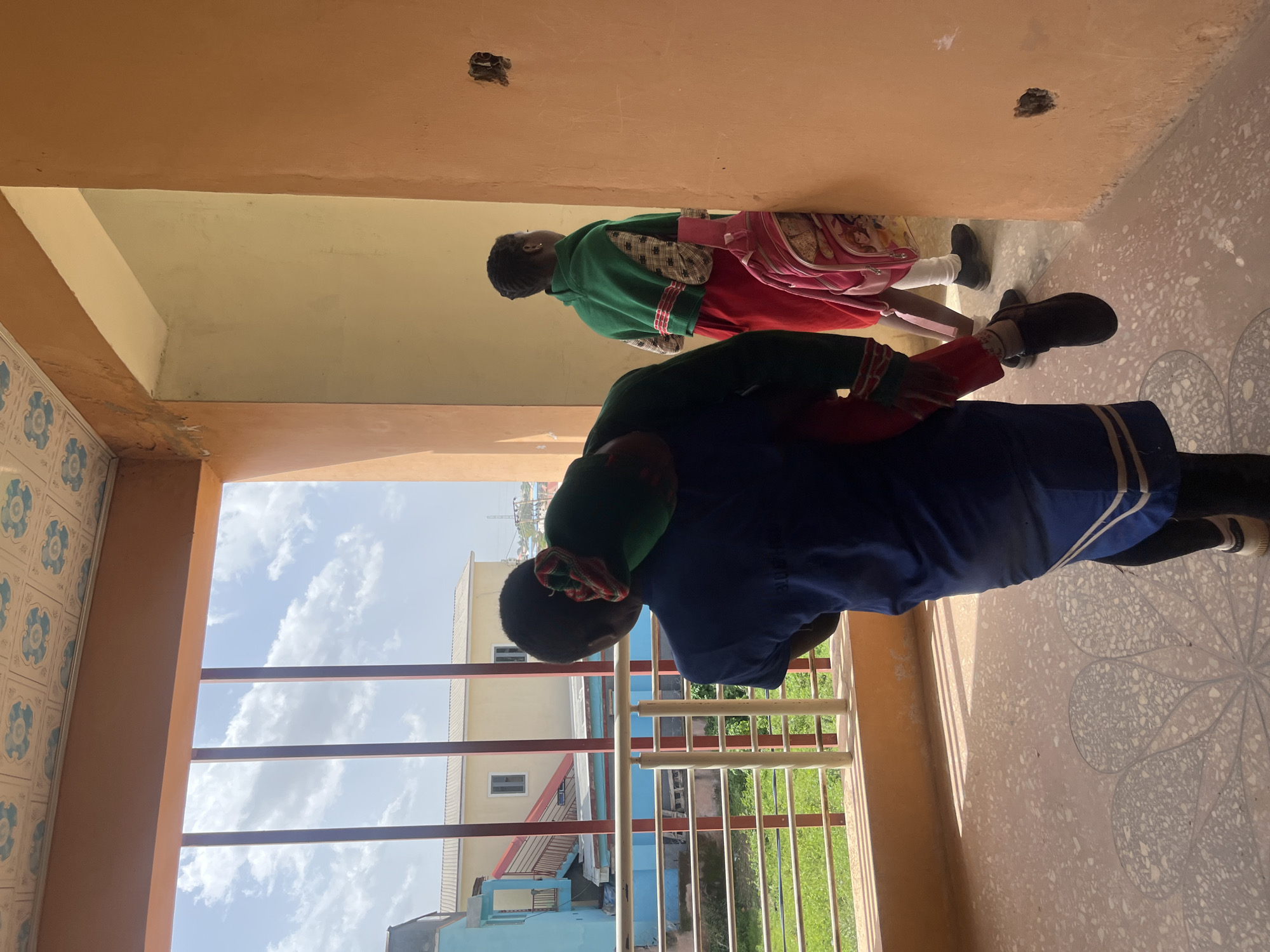 Two children are being carried by an older child, or adult, outside a building in Gura Top, Nigeria. The person carrying the two children is wearing a blue garment. The child in the middle is wearing a red garment and a green hooded jacket. The child on top is wearing a green hooded jacket. One of the children has a pink backpack with images of cartoon characters. The sun is shining, casting shadows on the floor. The location appears to be a residential area with buildings and greenery visible in the background. The sky is blue with some clouds. The overall impression is a warm, sunny day.